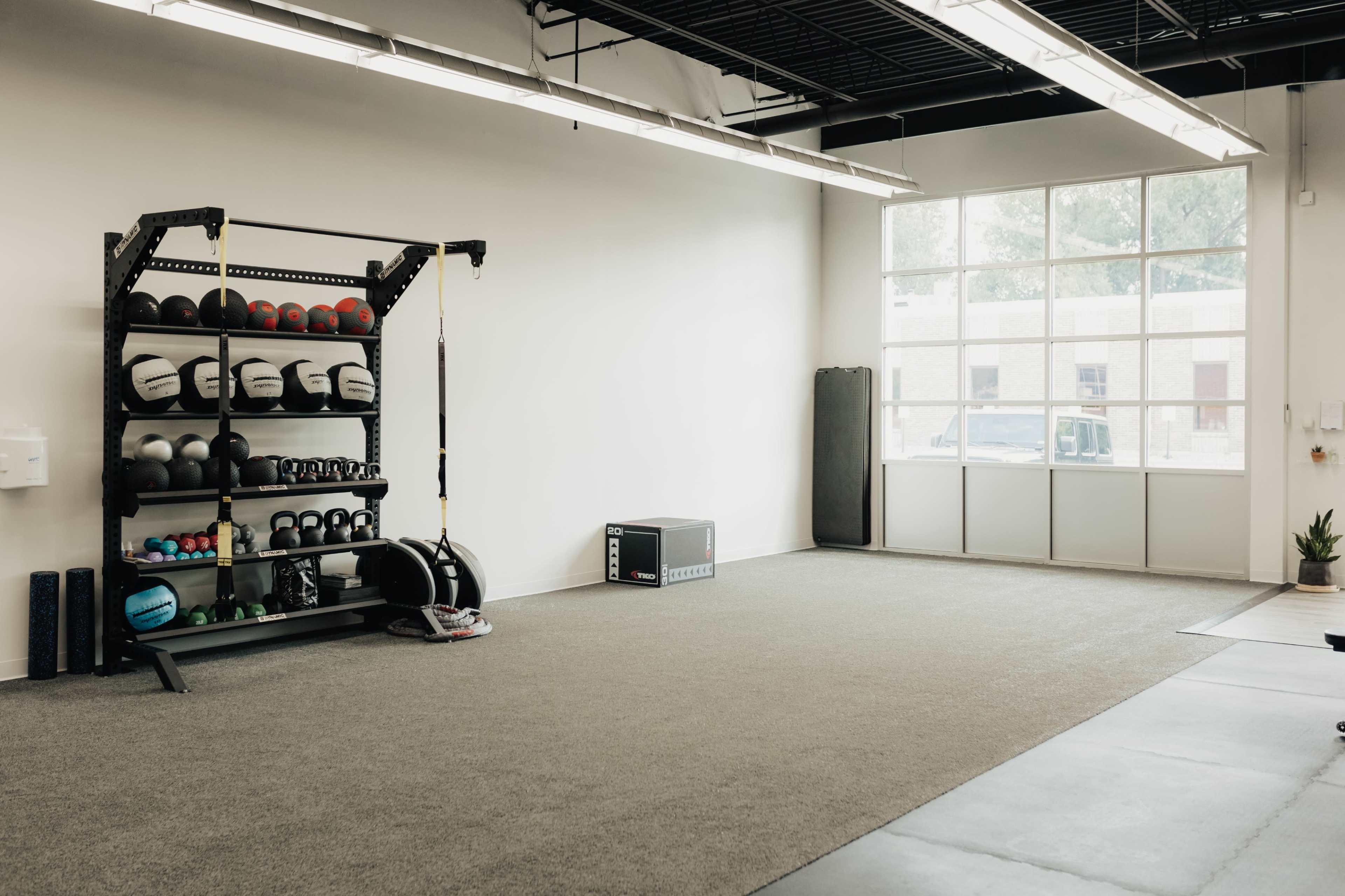 A spacious gym interior with a storage rack filled with various exercise equipment, large windows, and a clean, open floor area.