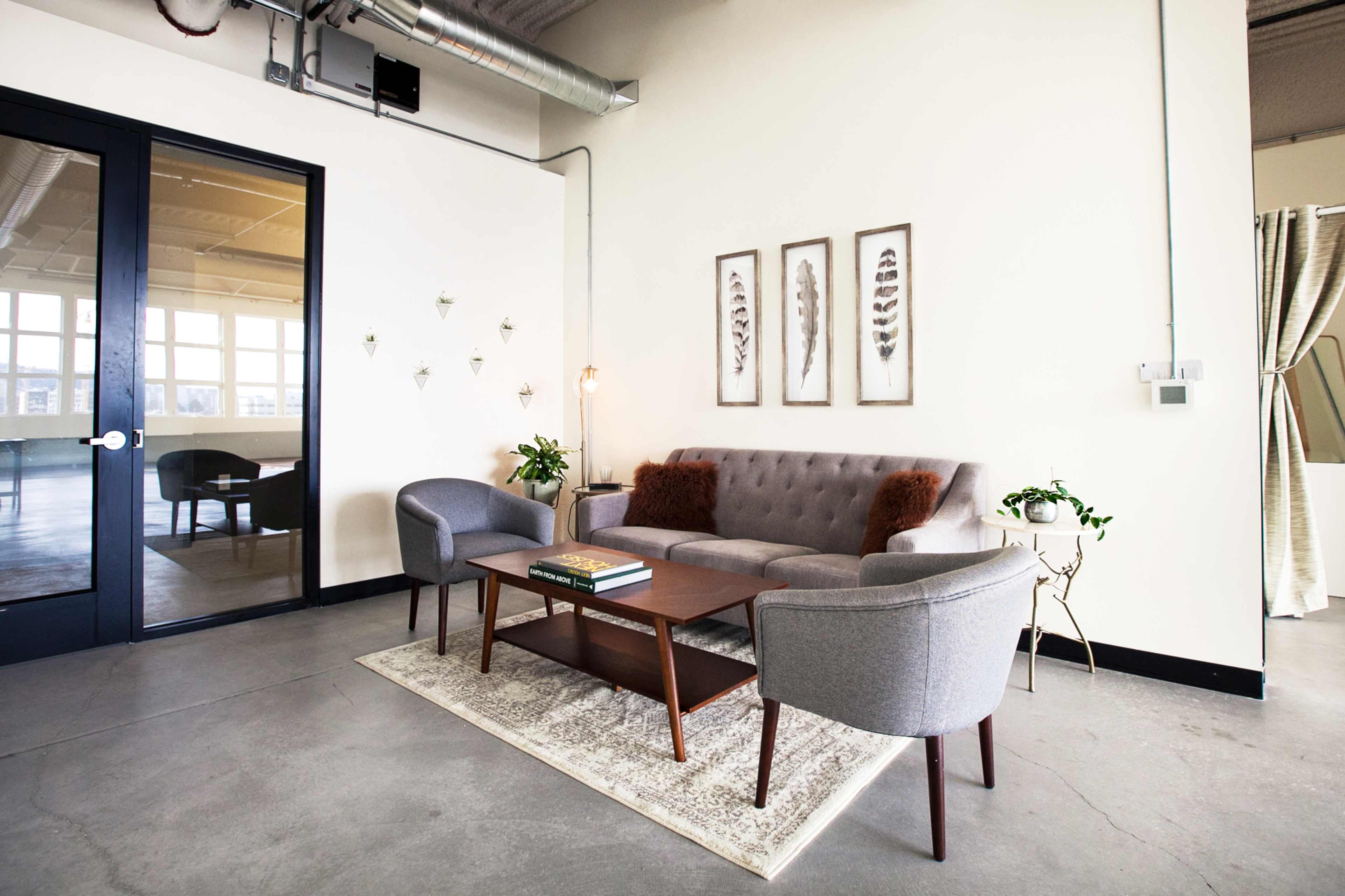 A modern, minimalistic lounge area features a gray sofa with decorative pillows, two matching armchairs, and a wooden coffee table, accented by framed artworks on the wall.