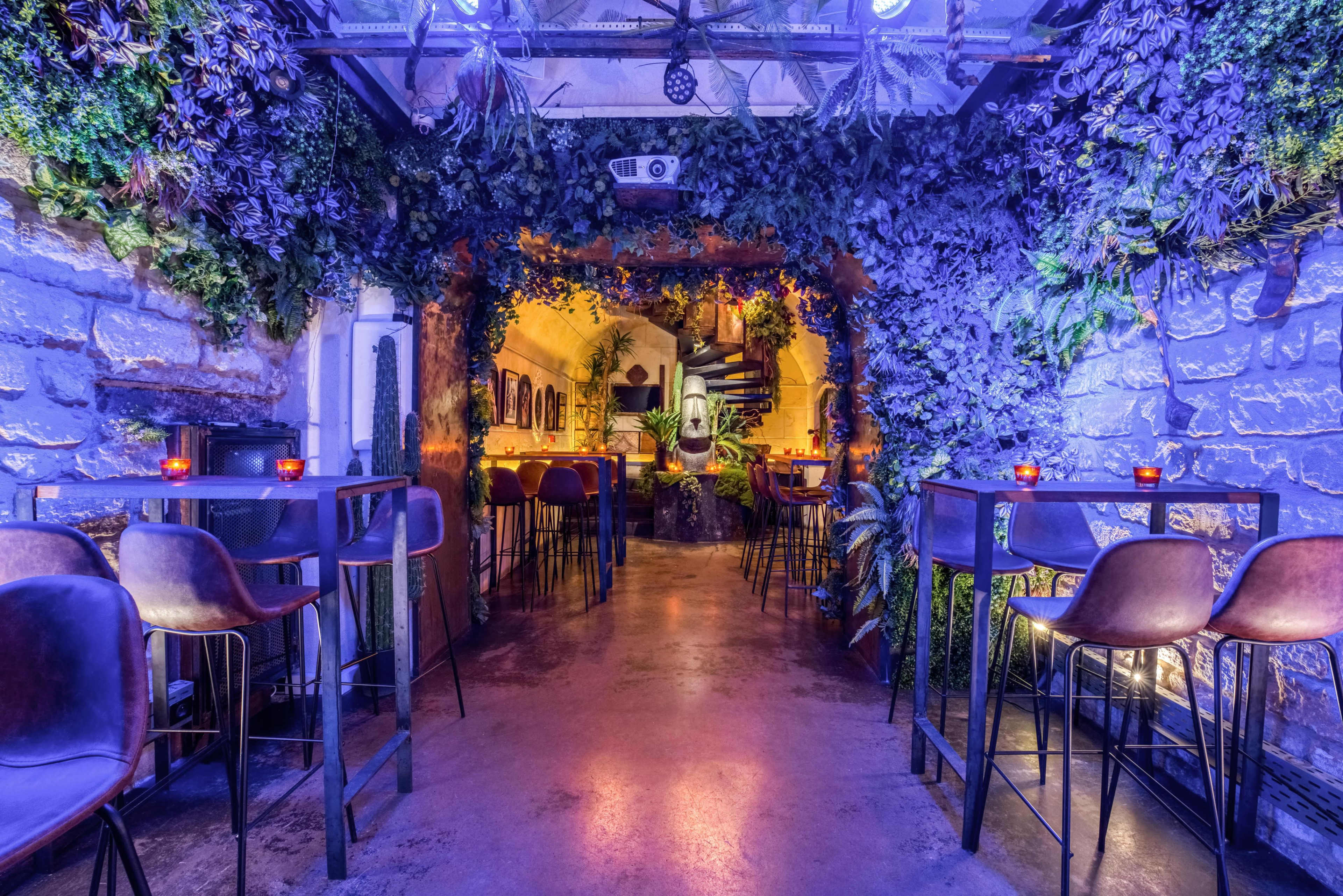 A dimly lit bar area with greenery-covered walls, small tables, and a staircase leading to another level.