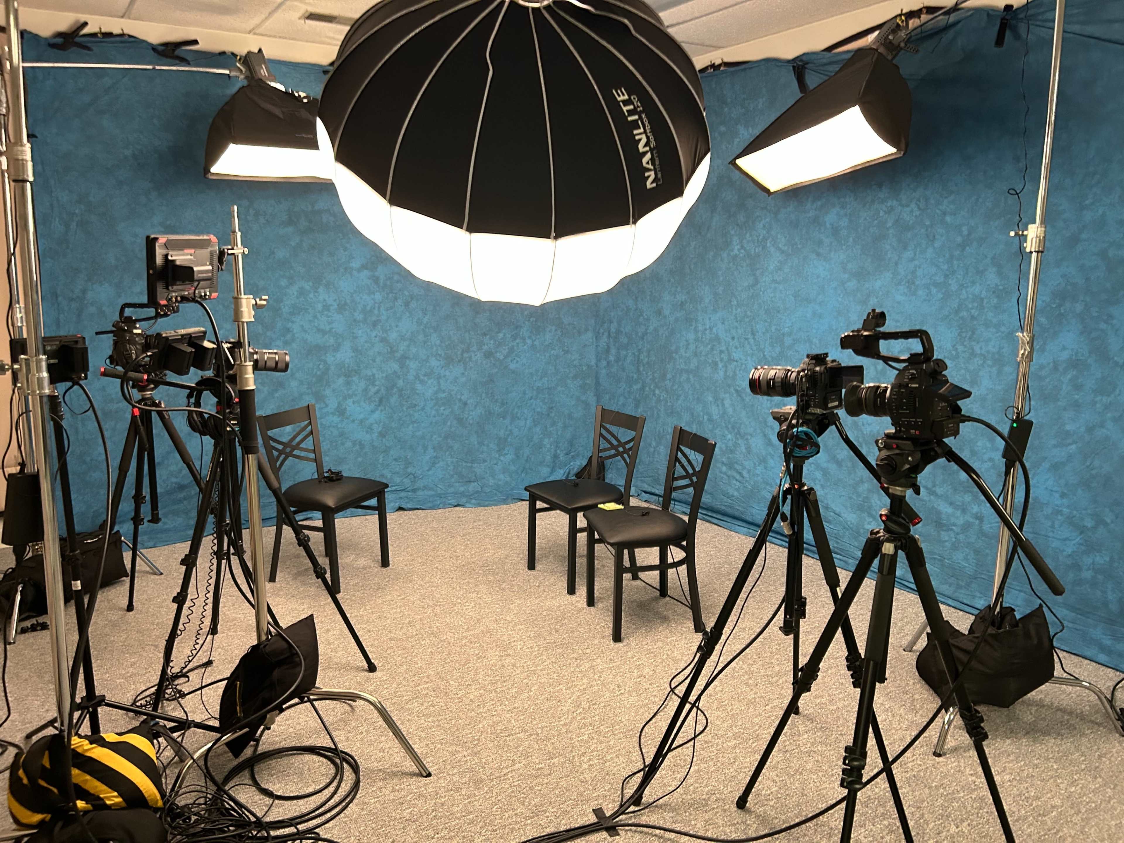 A photography studio is set up with three cameras and lighting equipment arranged around three empty chairs against a blue backdrop.