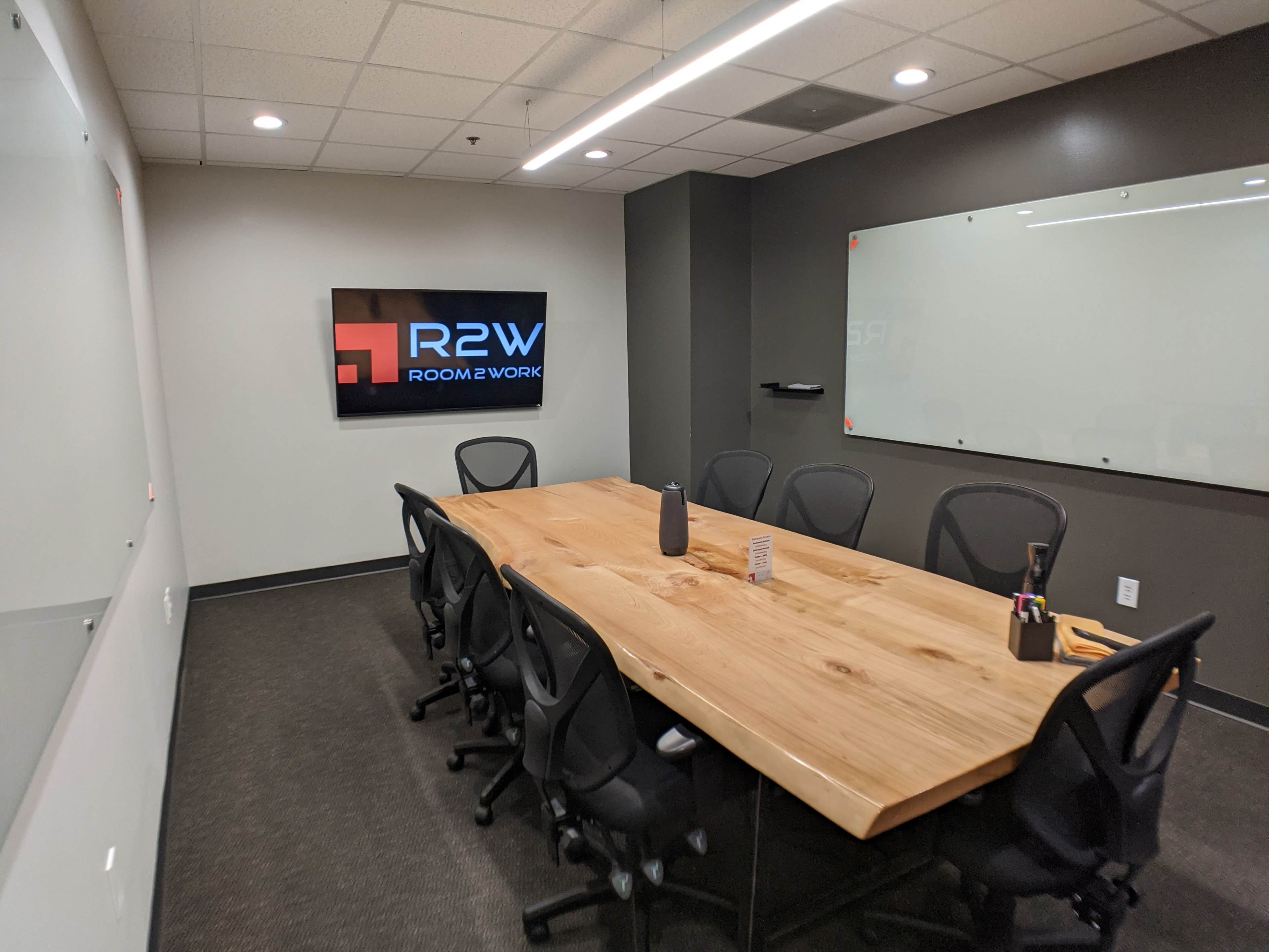 A modern conference room with a large wooden table surrounded by black chairs, featuring a wall-mounted screen displaying the "R2W Room 2 Work" logo.
