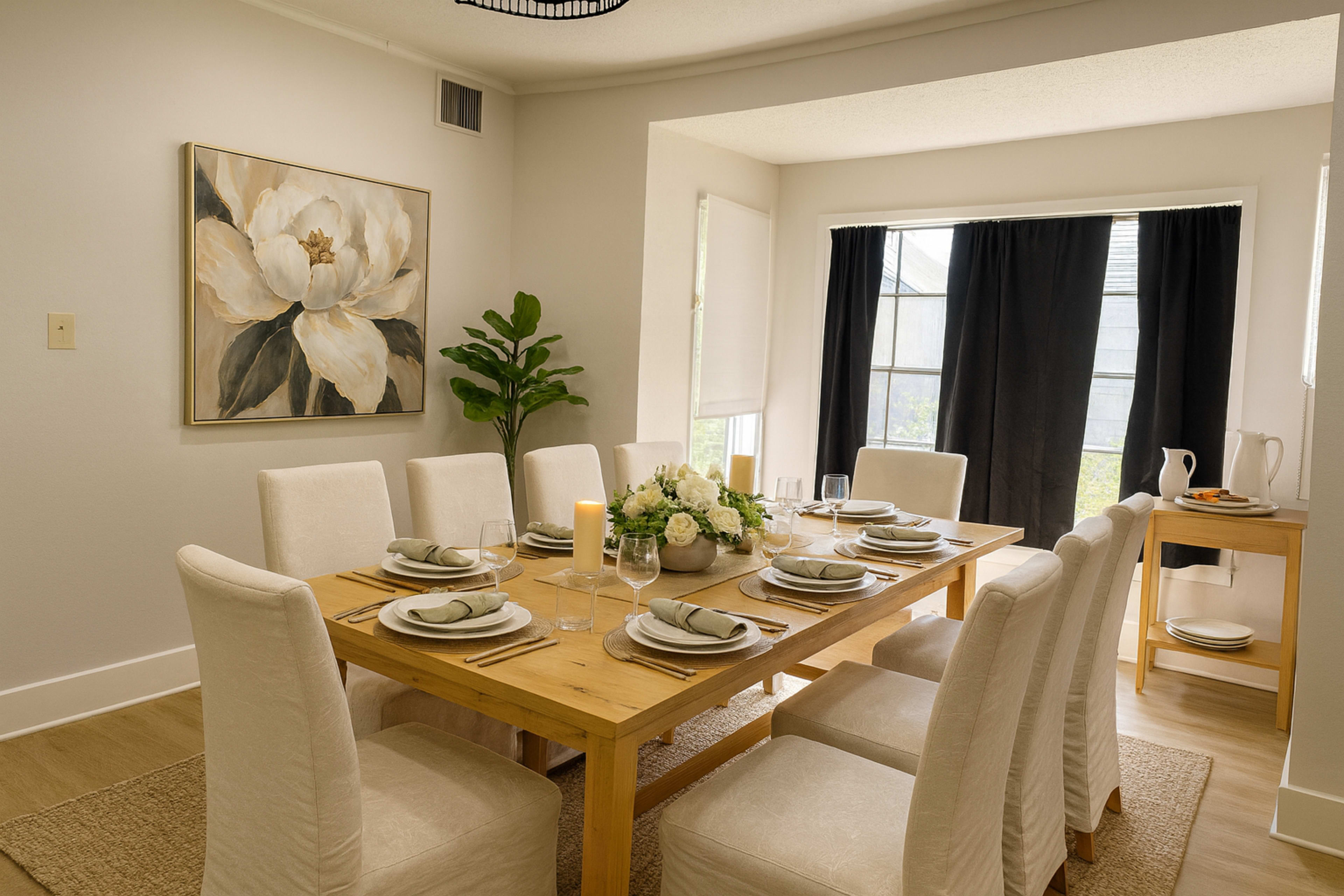 A dining room features a long wooden table set for eight, surrounded by white upholstered chairs, with a floral centerpiece and a large window dressed in black curtains.