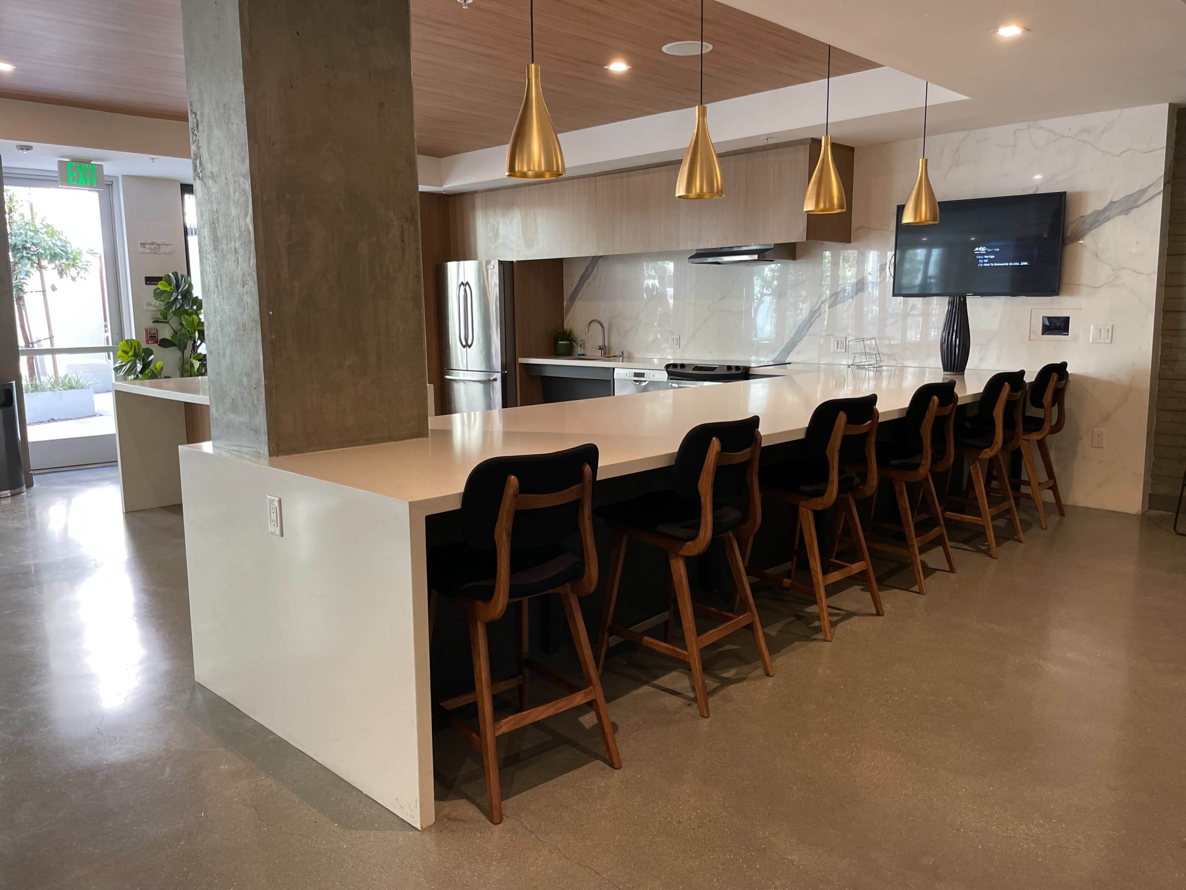 A modern kitchen with a long island bar and eight wooden stools, featuring pendant lighting and a large window overlooking a greenery-filled area.