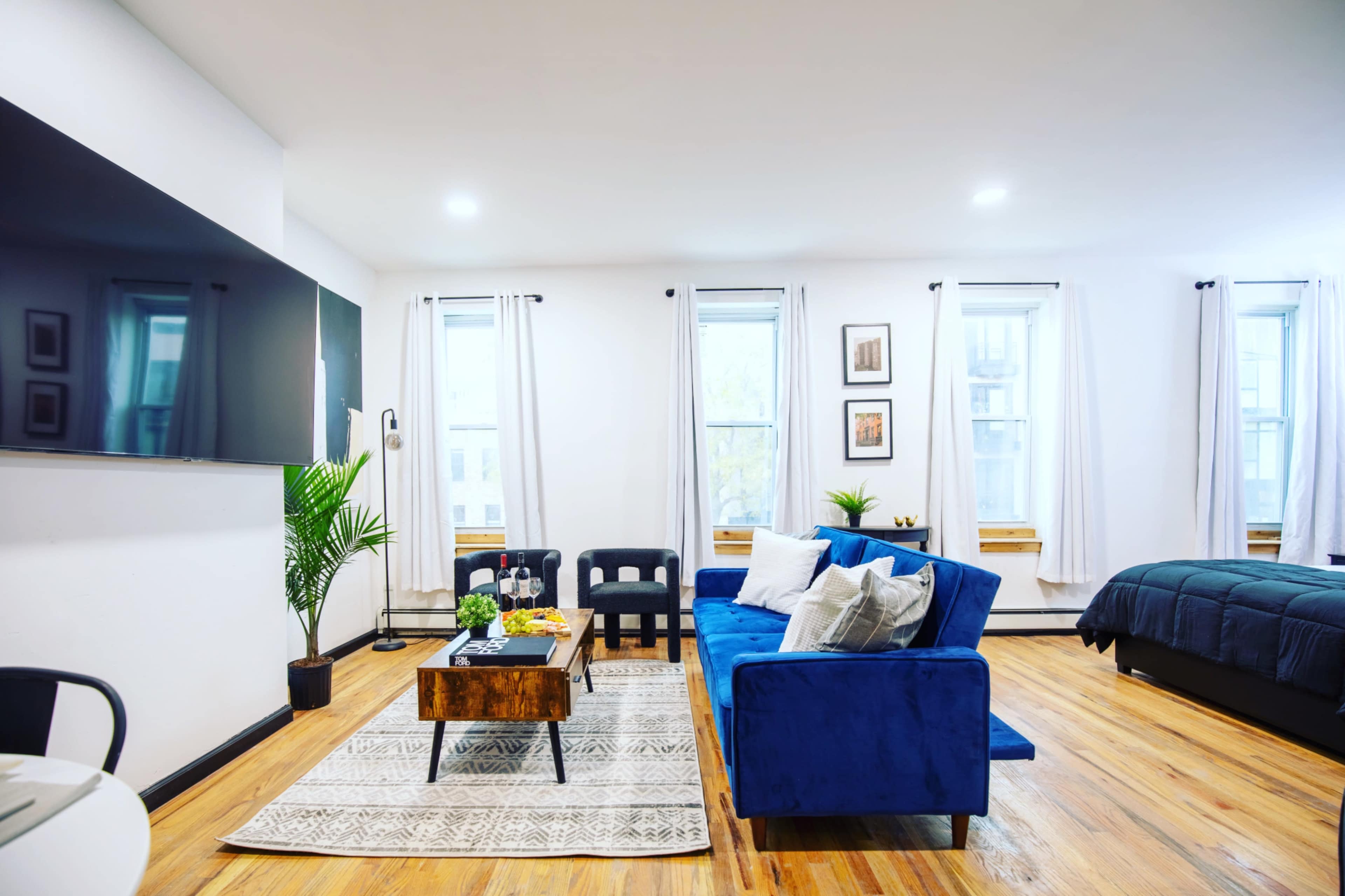 A modern living room features a blue sofa, a wooden coffee table, and a large television mounted on the wall, with windows allowing natural light to fill the space.