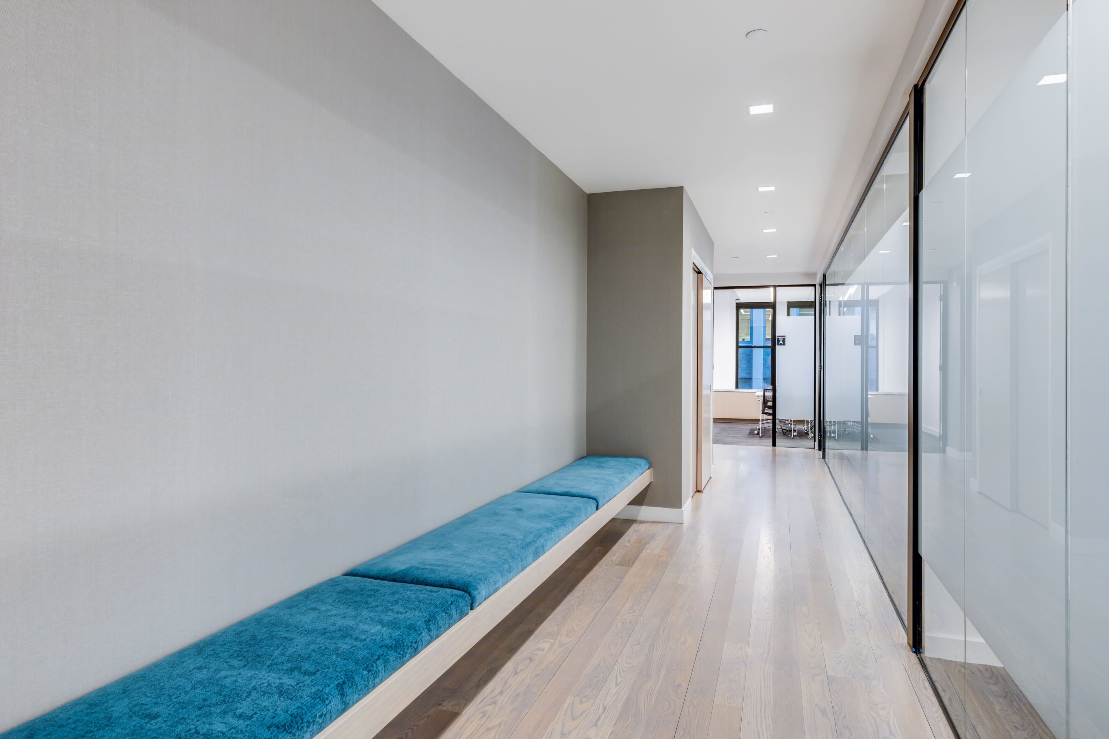 A narrow hallway with a light gray wall, a wooden floor, and a long blue bench along one side, with glass walls on the opposite side.
