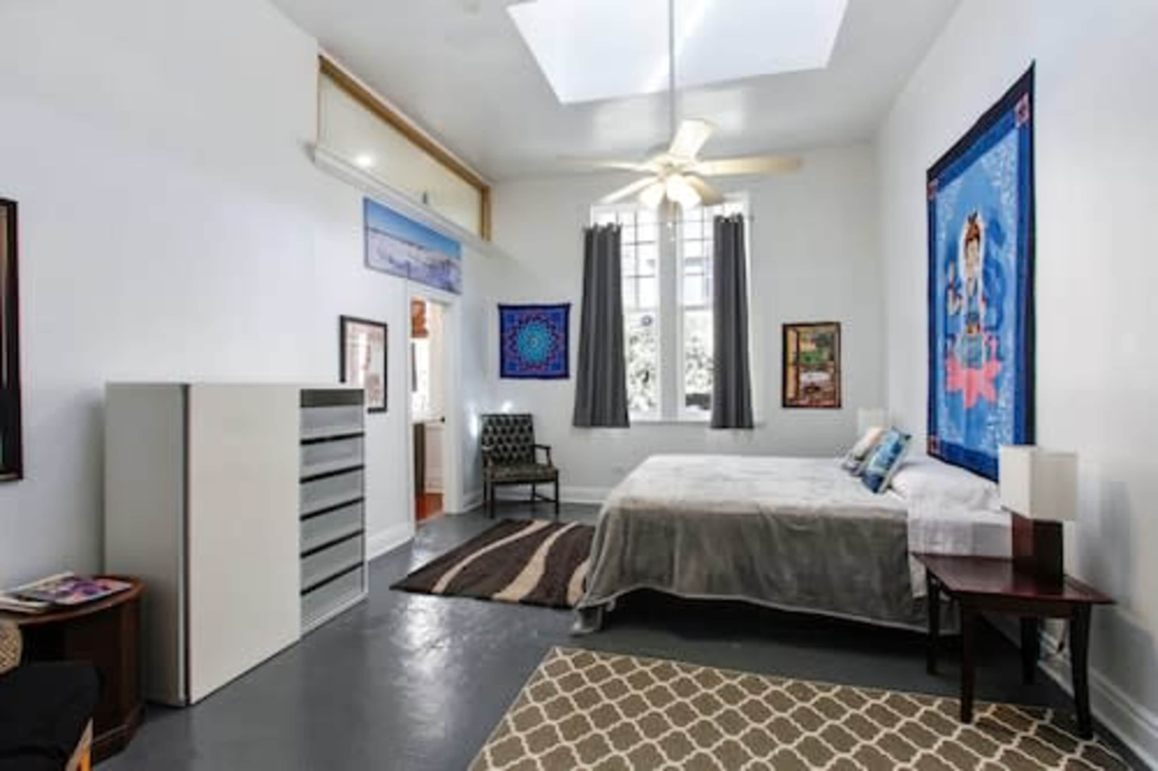 The image shows a spacious bedroom featuring a bed, a dresser, a ceiling fan, and large windows with curtains.
