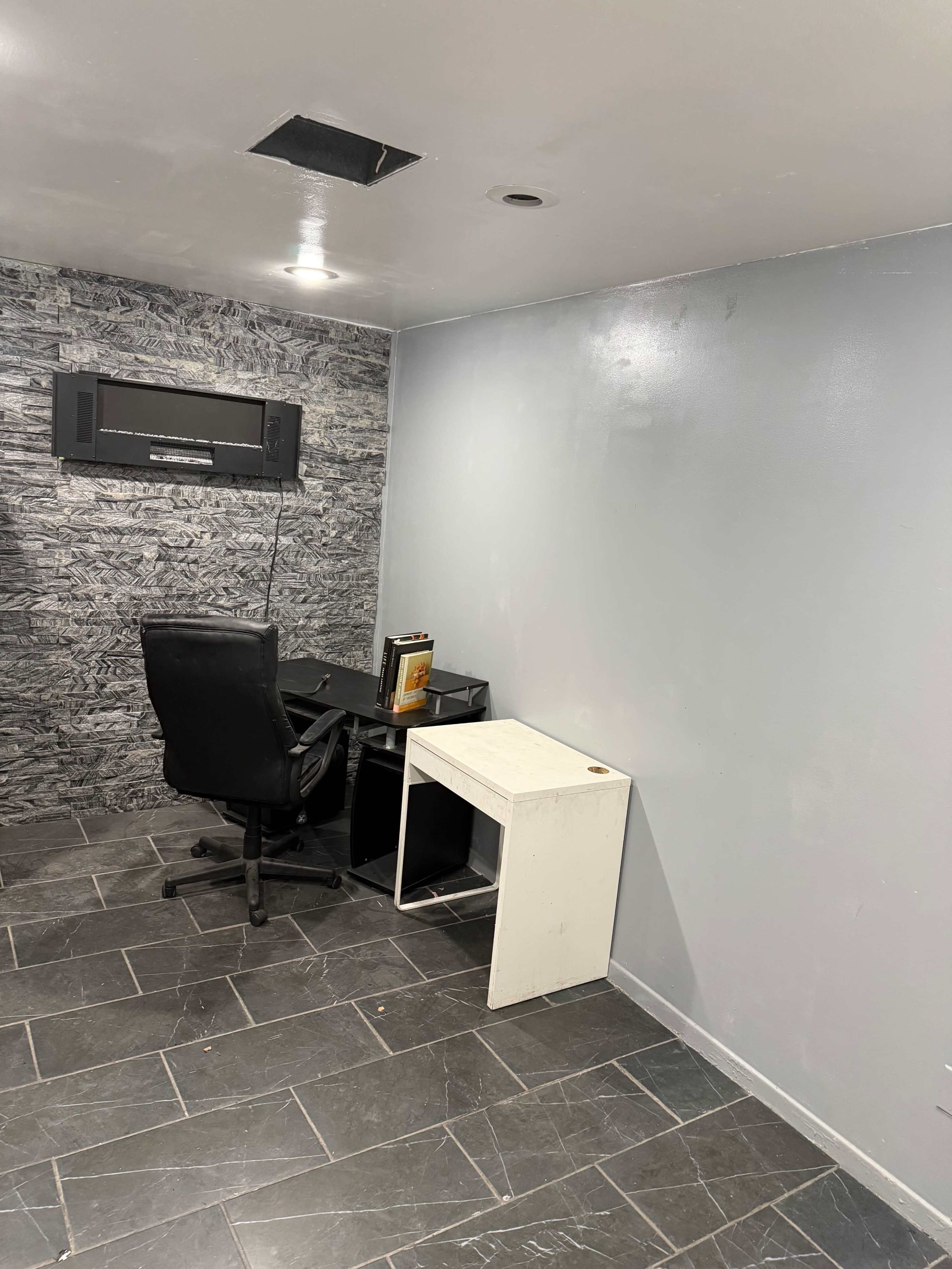 The image shows a sparse basement room with a black office chair, a desk against one wall, and a small white table next to it, along with patterned wallpaper and tile flooring.