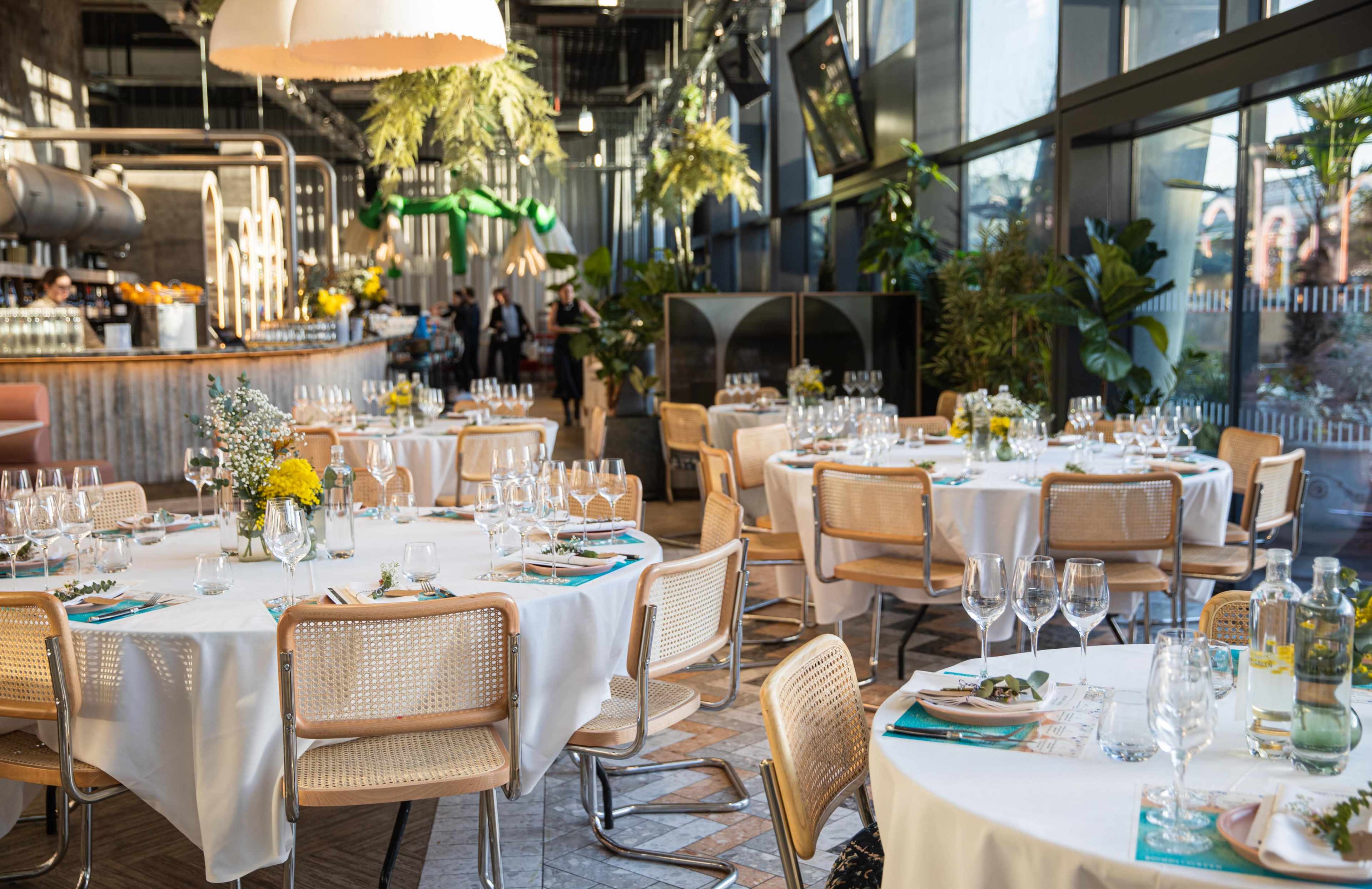 The image shows a modern dining area with round tables set for a meal, featuring white tablecloths, glassware, and floral centerpieces, surrounded by large windows and greenery.