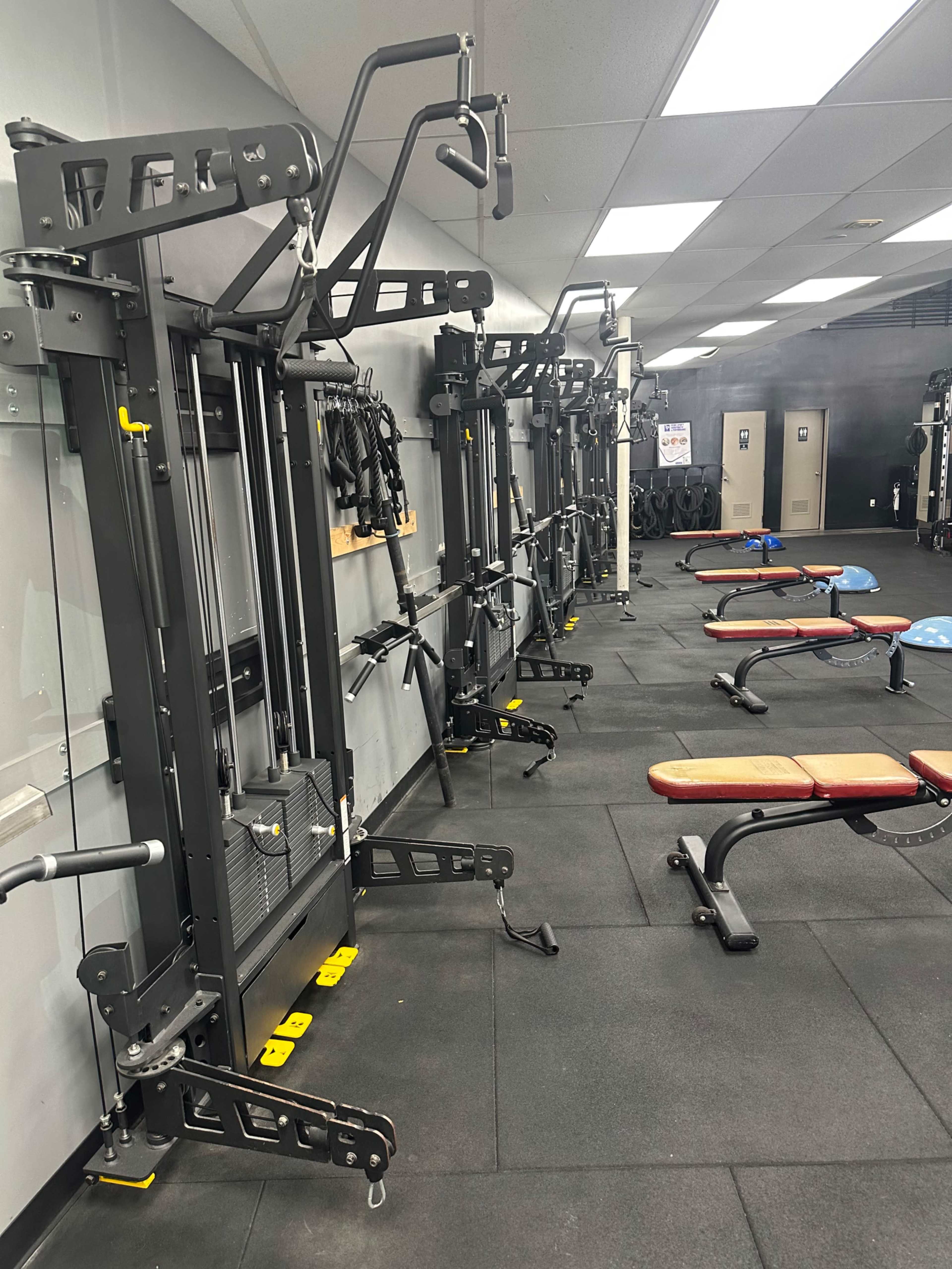 The image shows a gym interior with multiple weight machines and benches arranged on a black rubber floor.