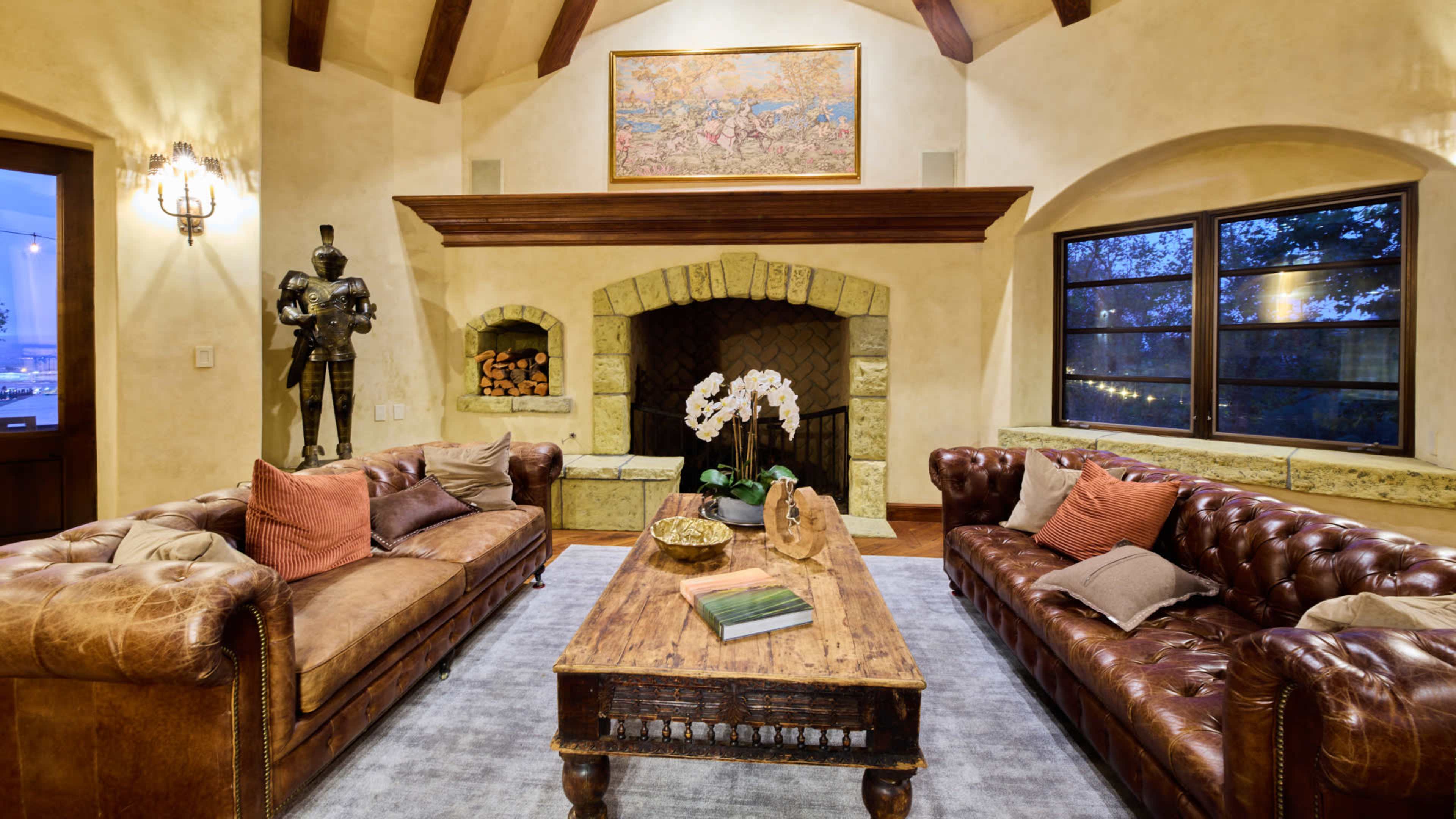 The living room features two brown leather sofas facing a wooden coffee table in front of a stone fireplace, with a decorative artwork hung above.
