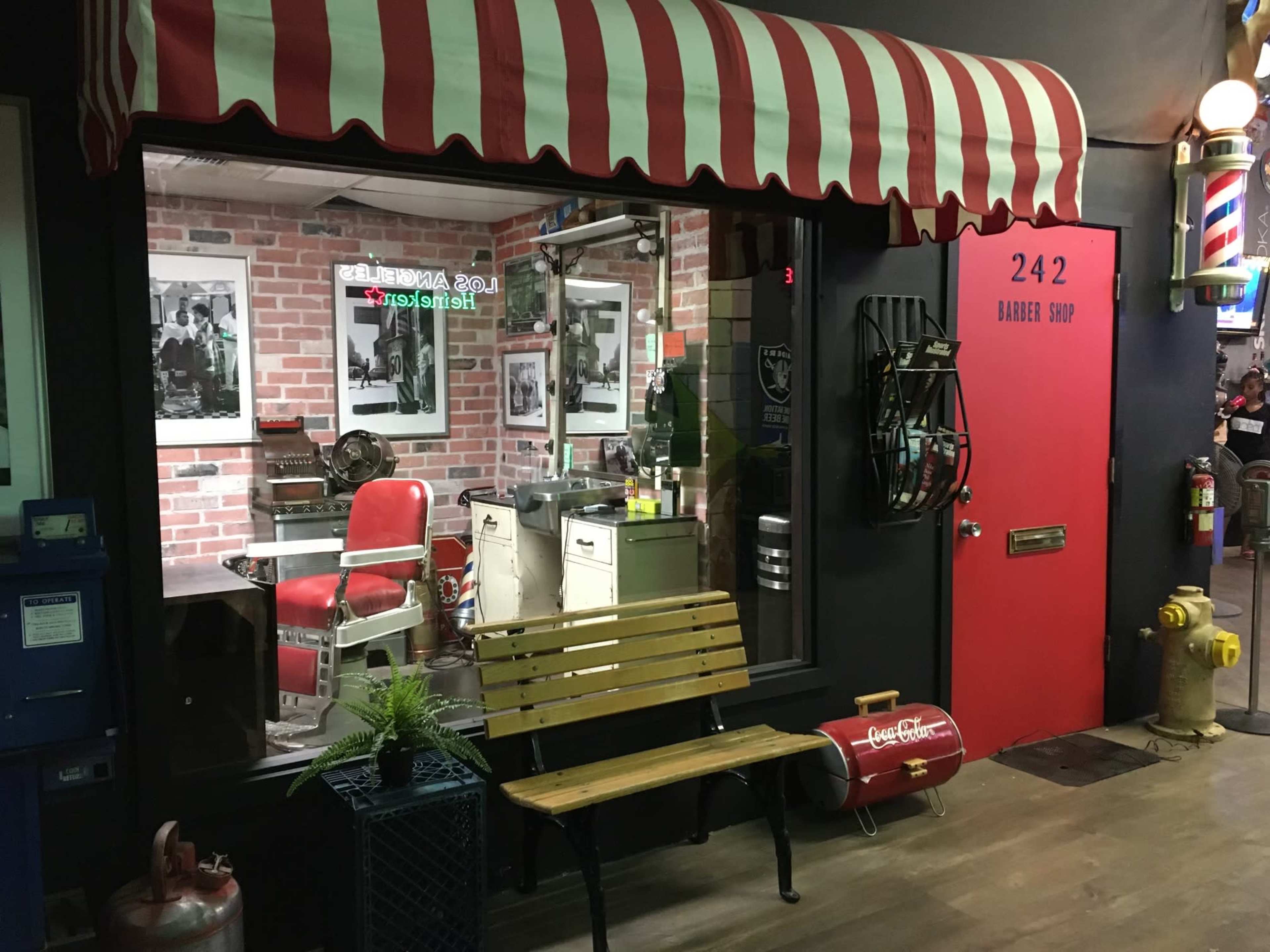 A vintage-style barber shop with a red and white striped awning, featuring a classic barber chair, antique equipment, and black-and-white photos on the brick walls.