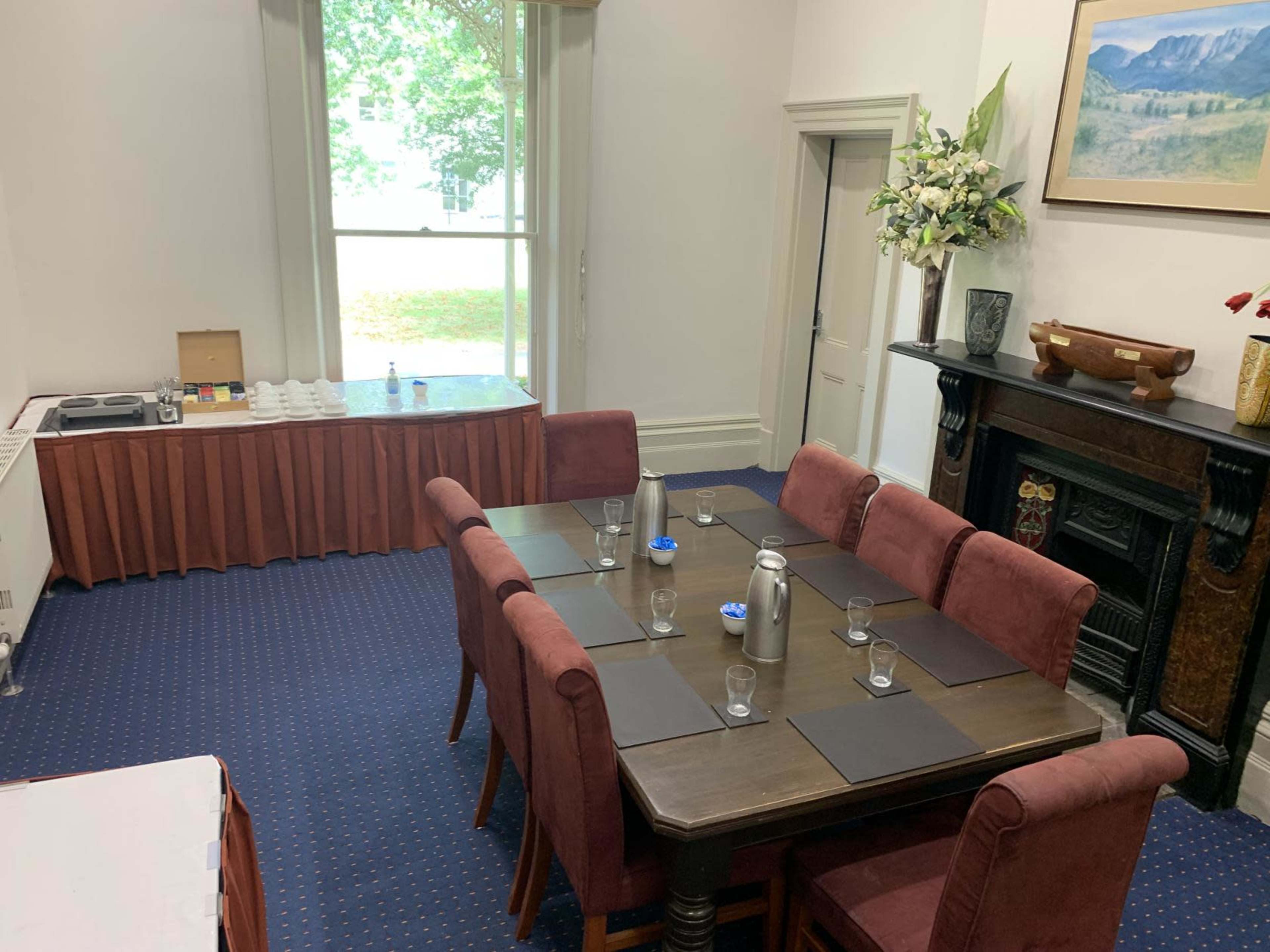 A dining area with a rectangular table set for a meal, surrounded by upholstered chairs, a side table with refreshments, and a large window providing natural light.