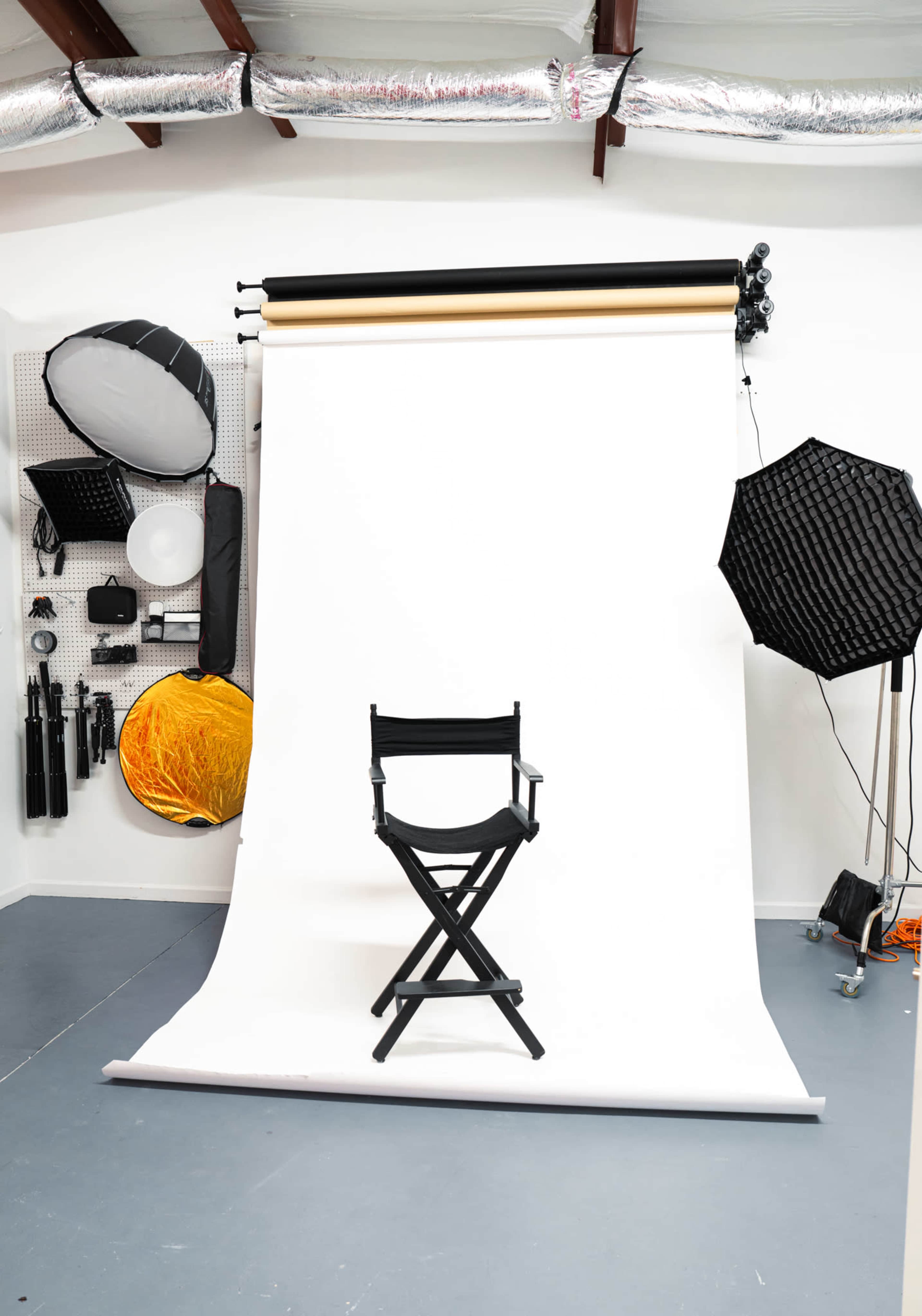 A wooden director's chair is placed in front of a large white backdrop in a photography studio, surrounded by various lighting equipment.