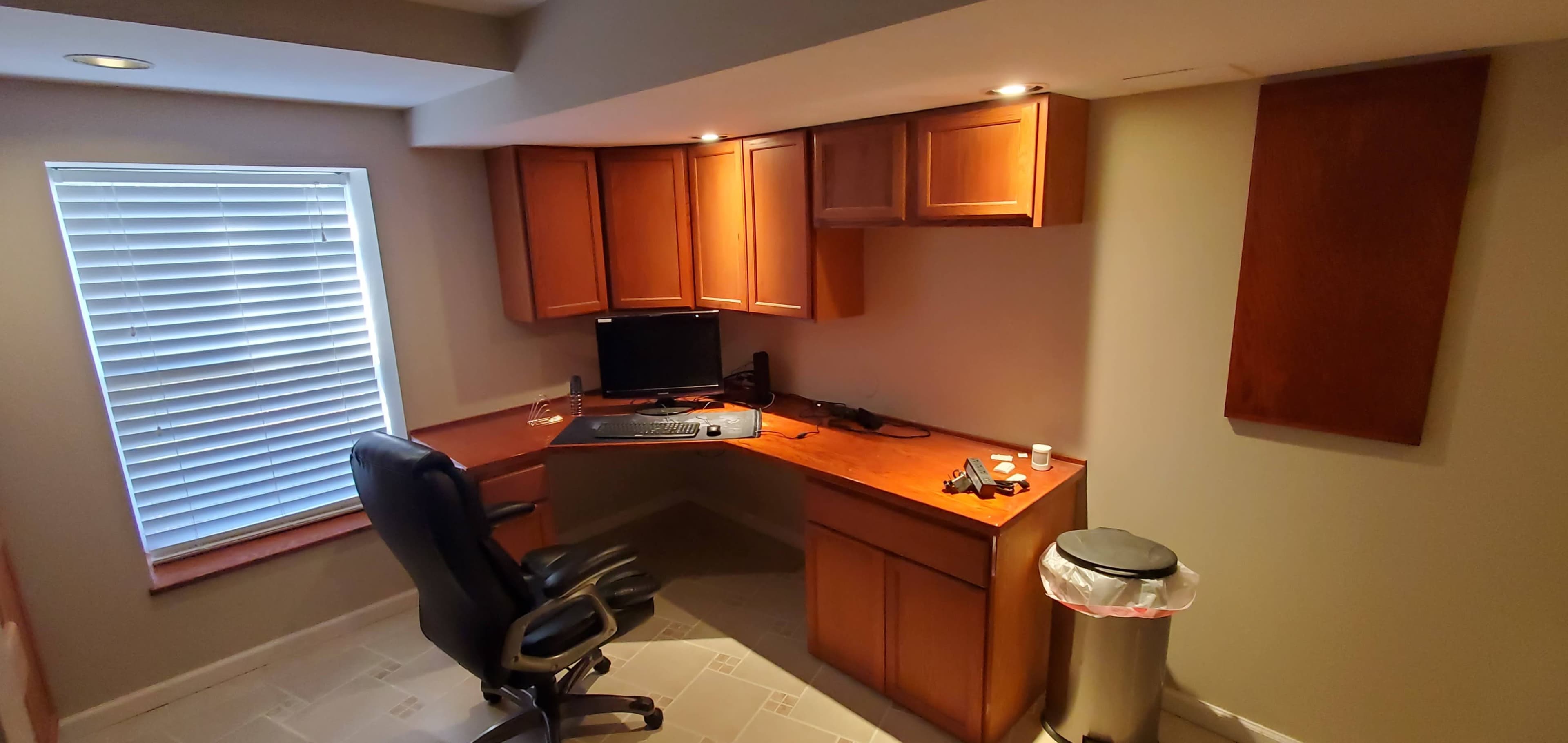 A wooden desk with a computer monitor, office chair, and trash can in a home office with a window featuring blinds.
