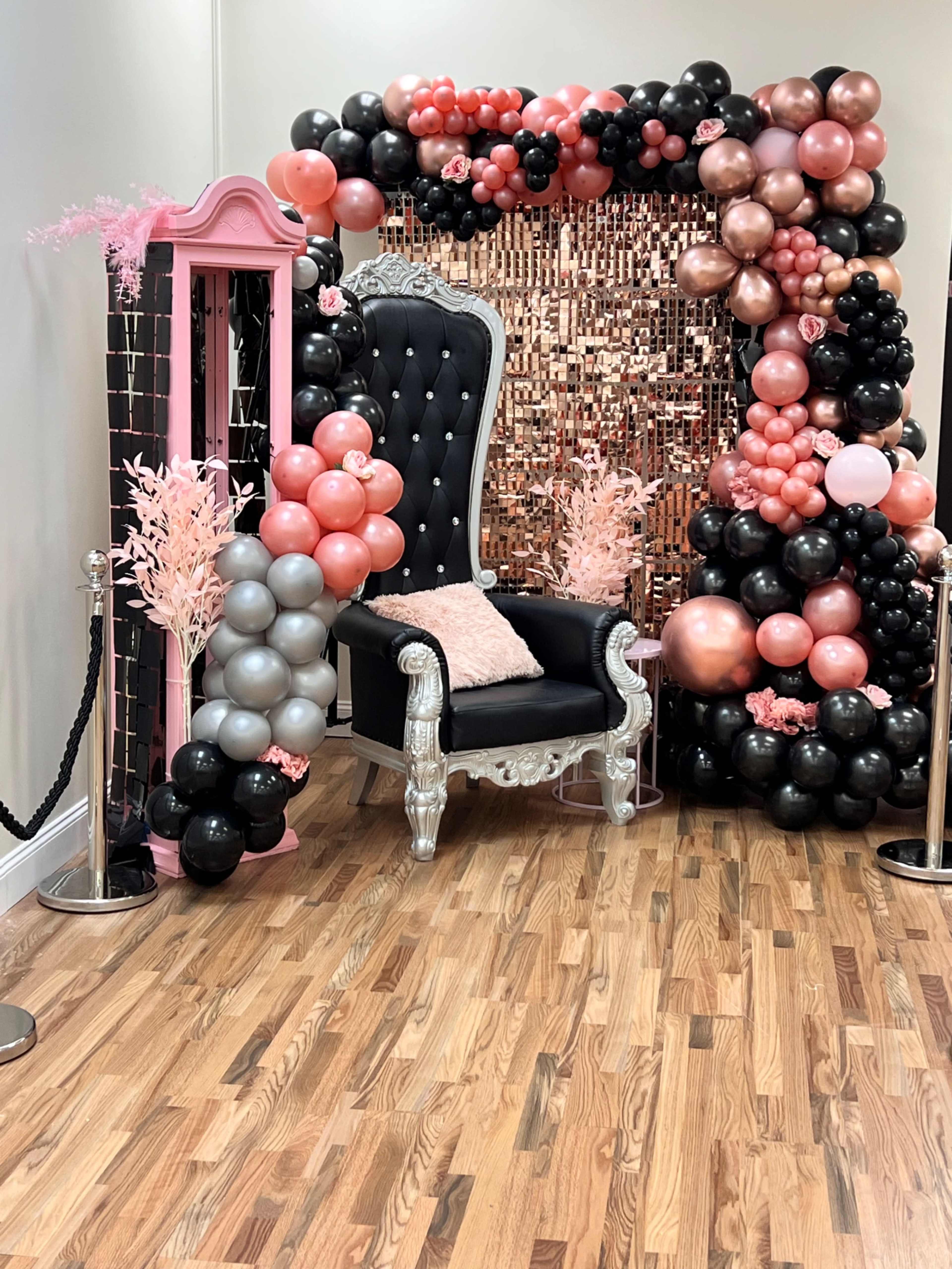 A decorative setup featuring a black throne chair surrounded by a balloon arch in pink, black, and silver, with a pink floral display and a mirrored wall backdrop.