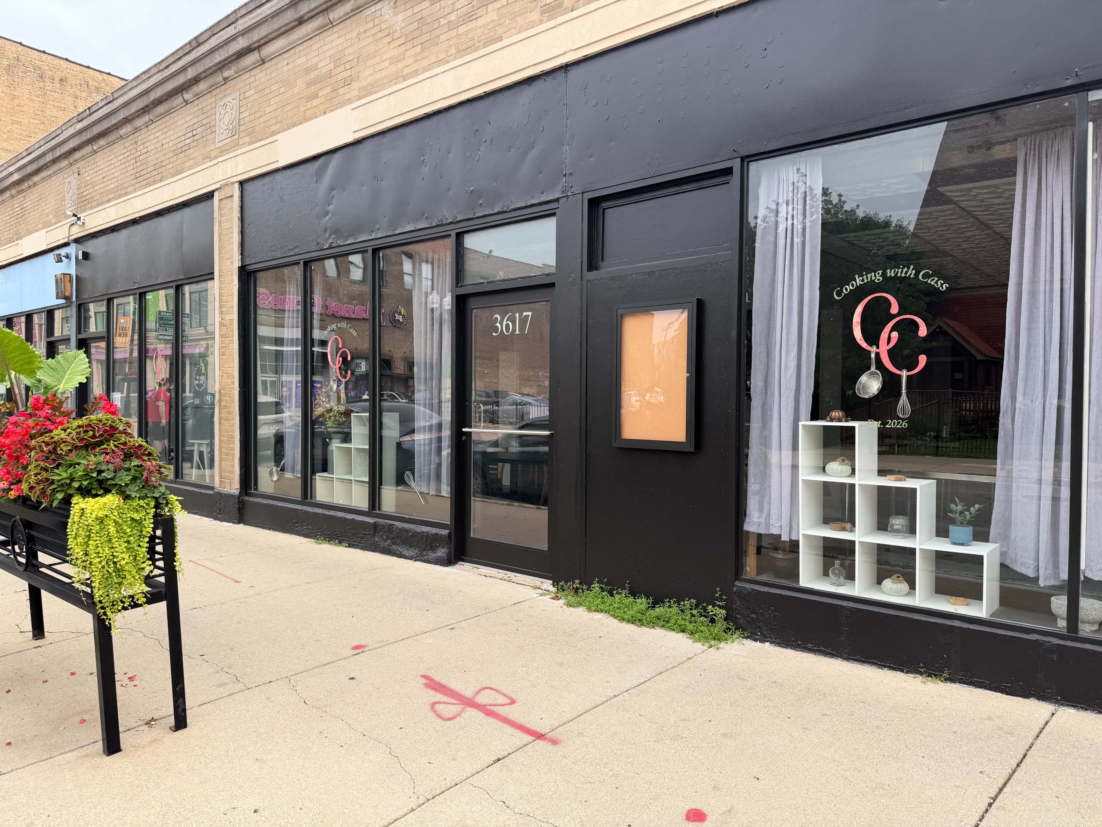The image shows a storefront with large windows, featuring a sign for "Cooking with Cake" at 3617, flanked by floral arrangements on the sidewalk.