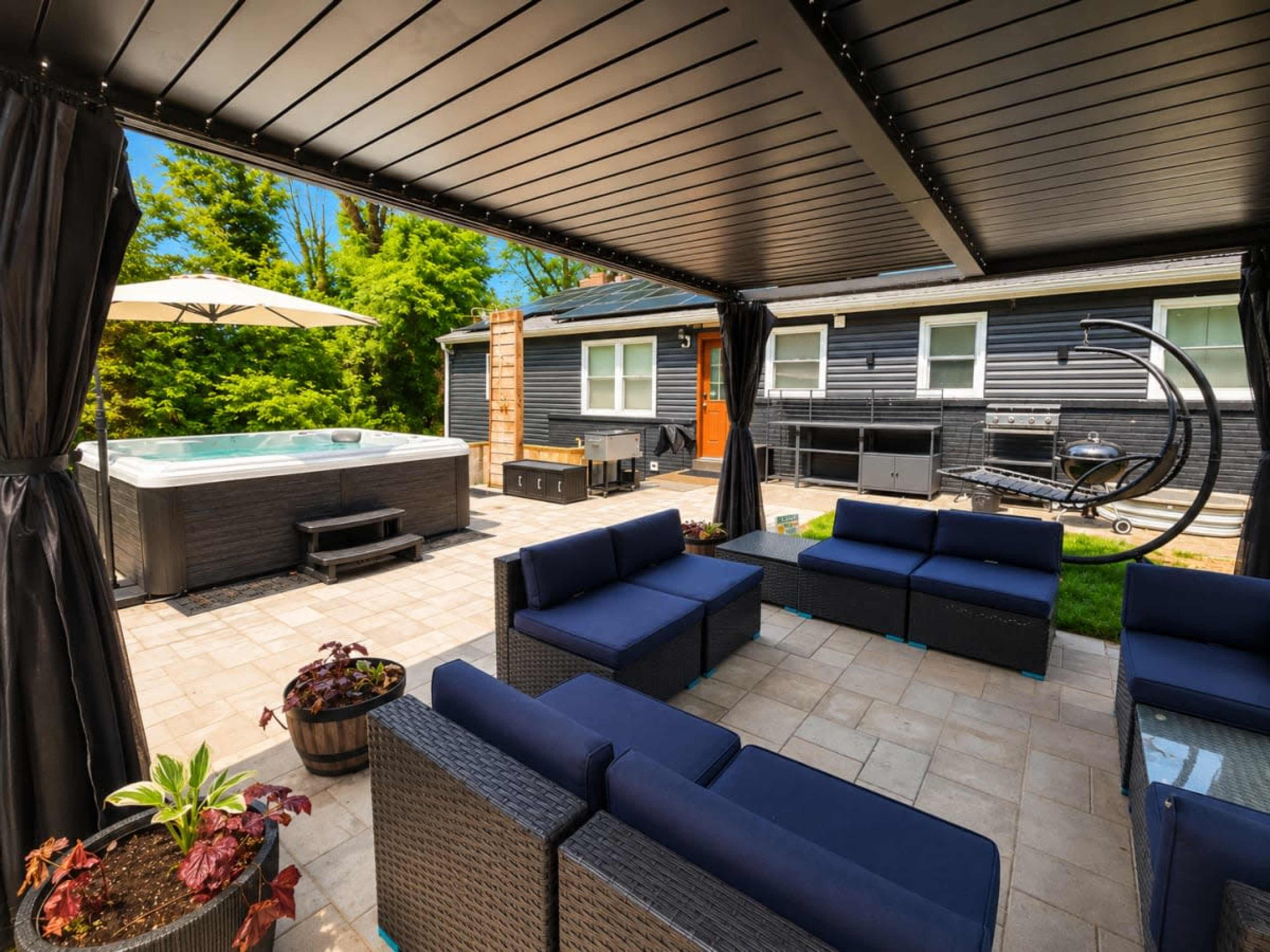 The image shows a stylish outdoor patio area with seating, a hot tub, and a grill, surrounded by greenery.