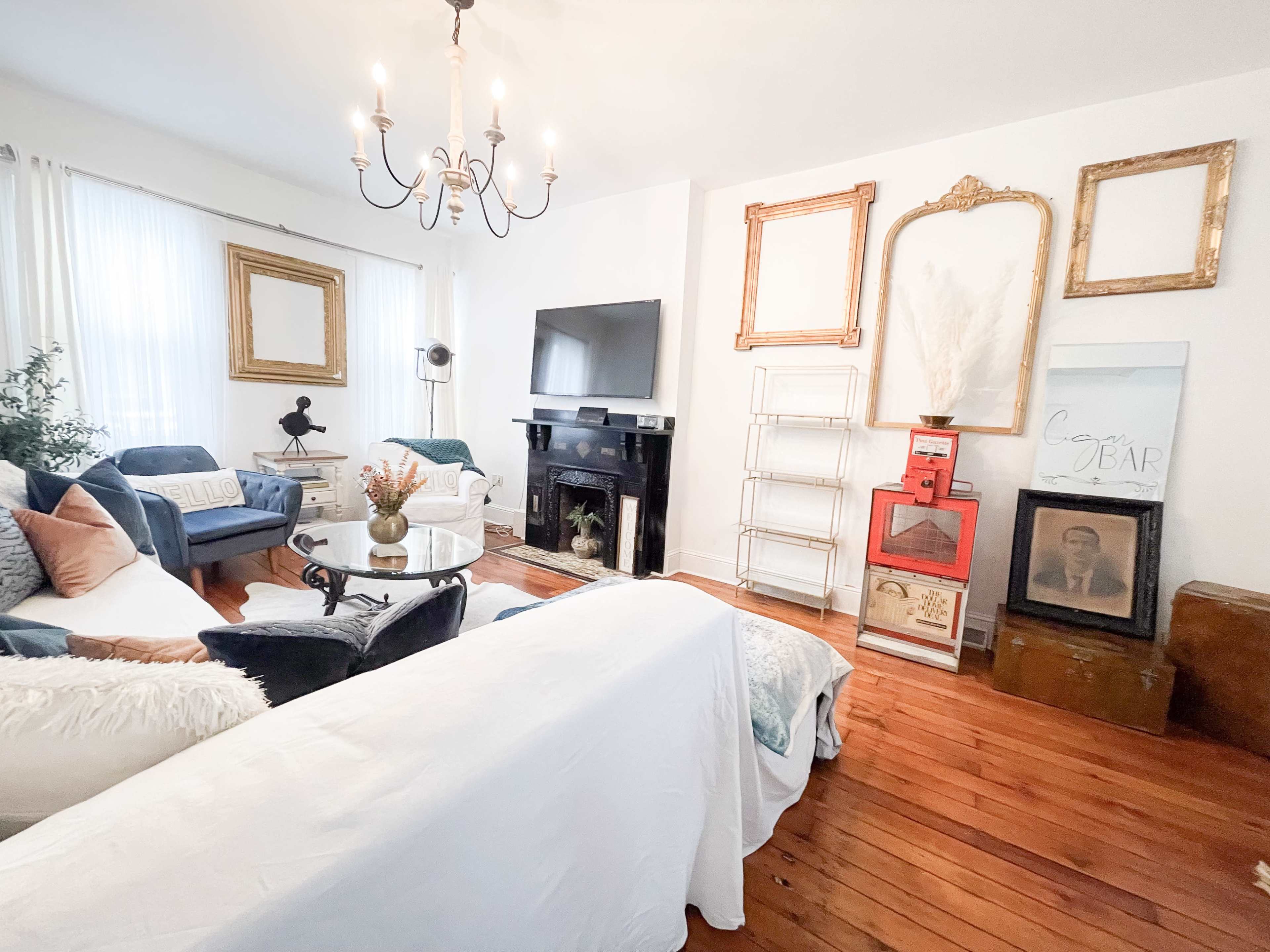 A cozy living room features a chandelier, a large TV mounted on the wall, and a mix of furniture including a white sofa and blue chairs, with decorative elements like frames and a vintage display cabinet.