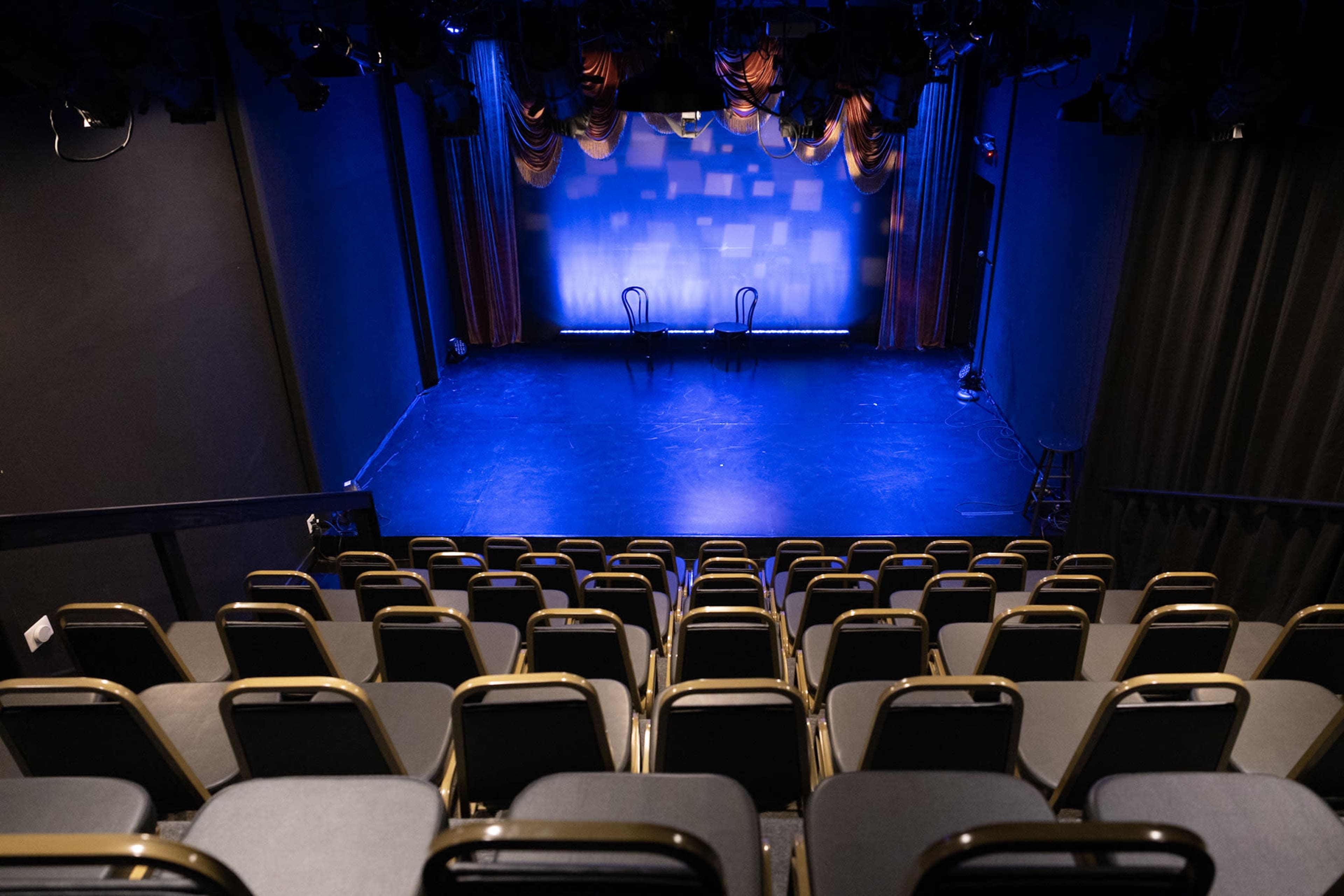 A small theater stage with two chairs and blue lighting, viewed from an elevated seating area filled with empty rows of chairs.