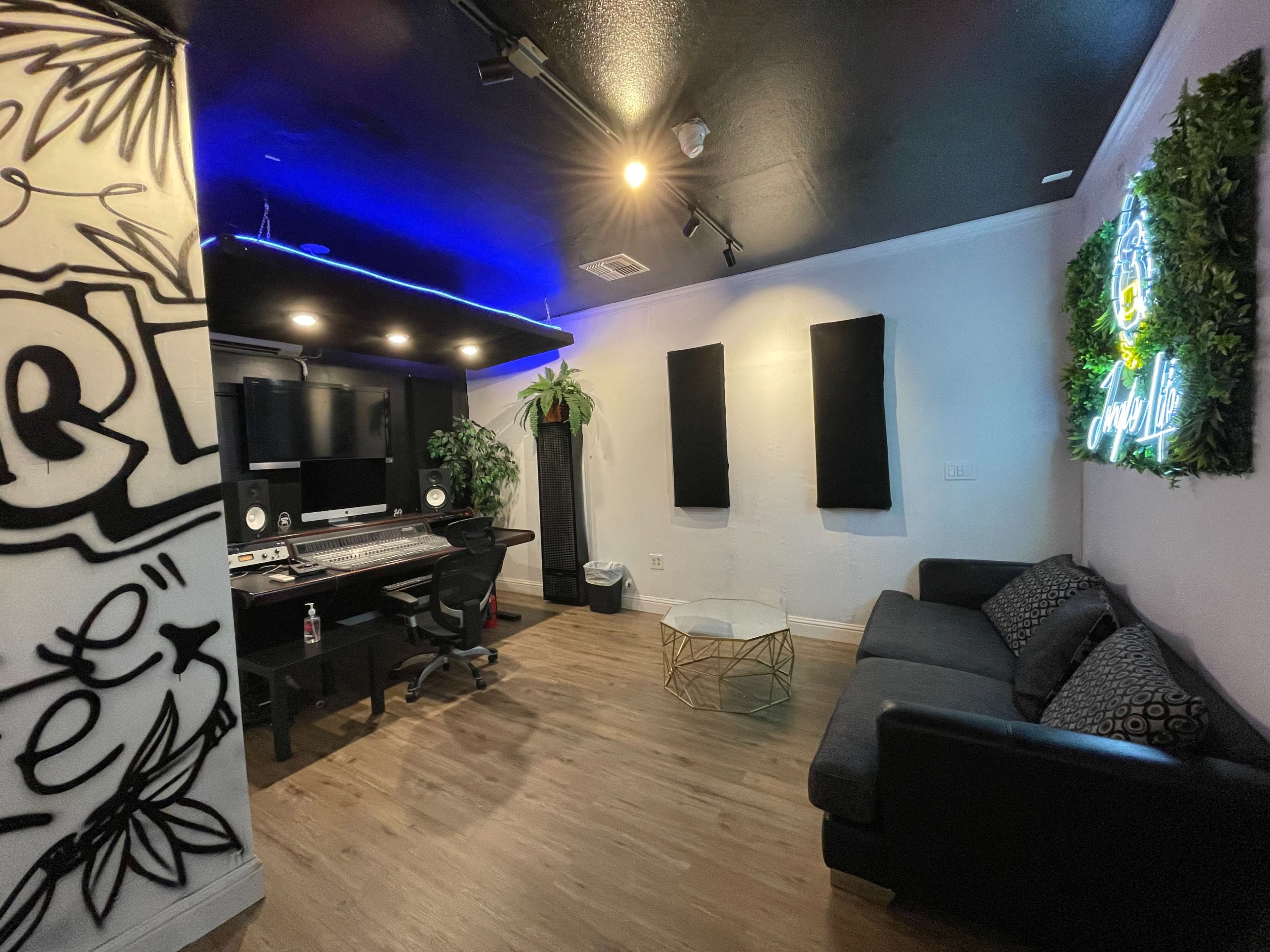 The image shows a music studio with a mixing desk, computer setup, black couch, and decorative plants.