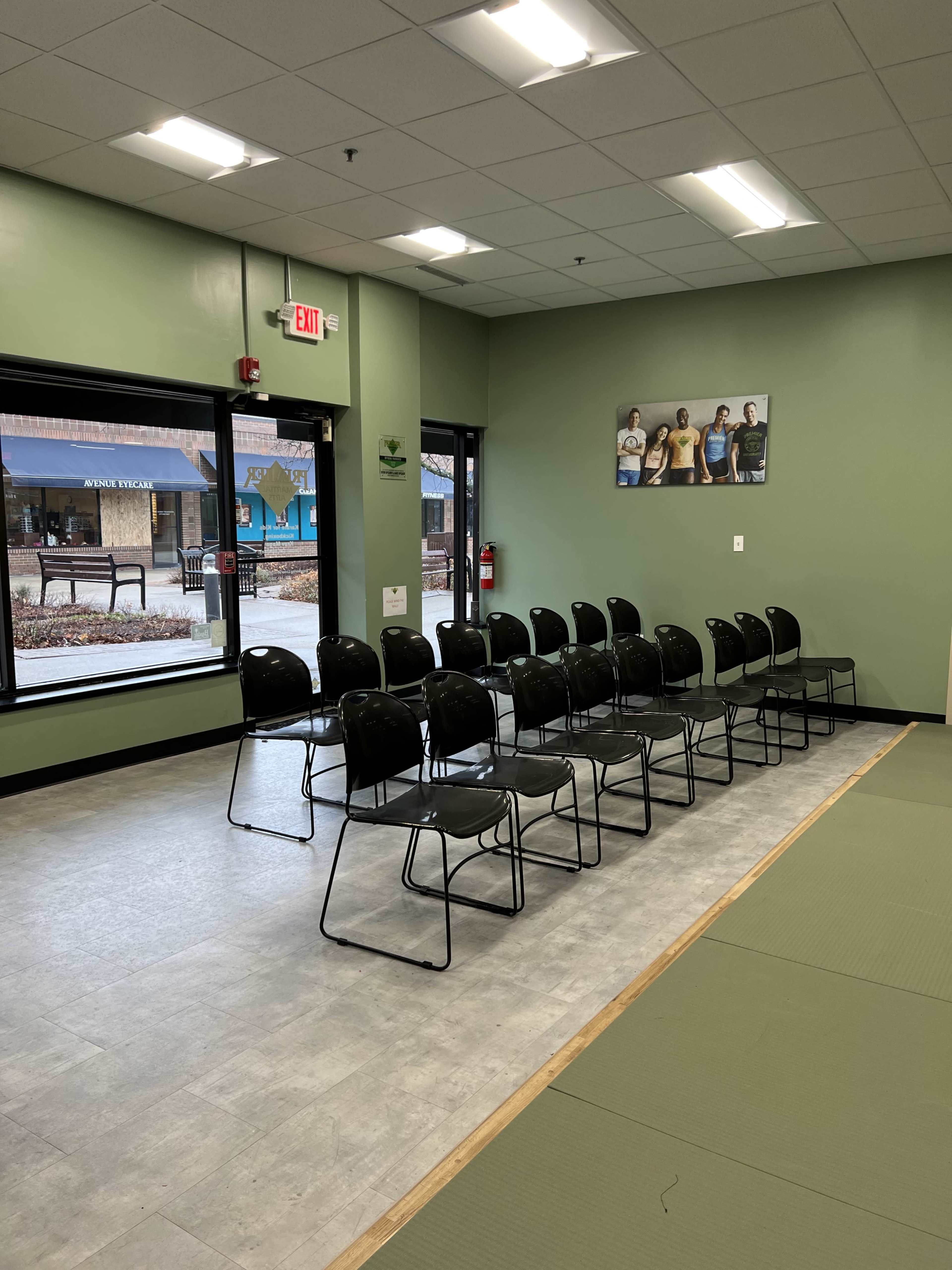 The image shows a row of black chairs lined up in a brightly lit room with green walls and large windows.