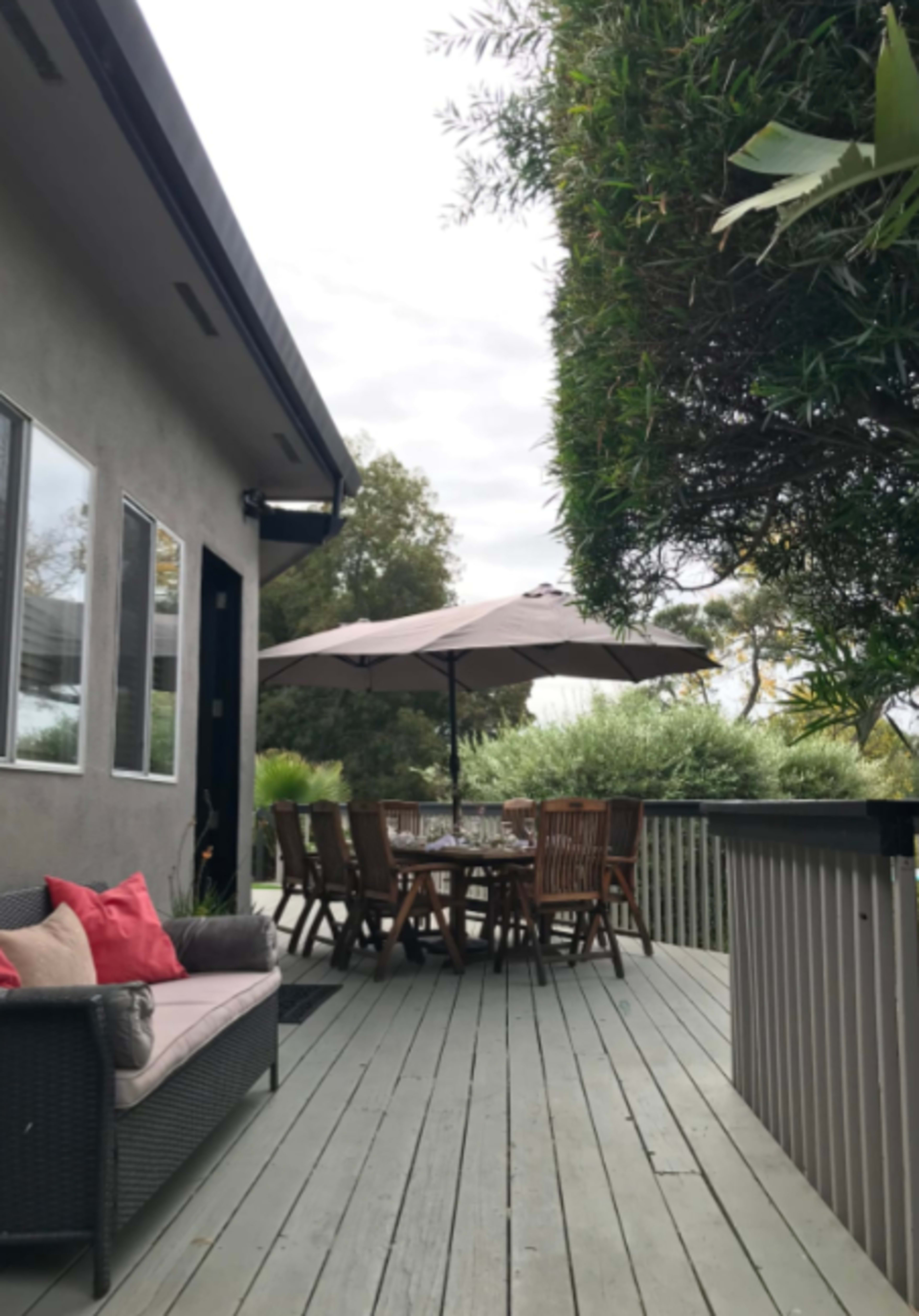 A wooden deck features a dining table with chairs, an umbrella, and a seating area, surrounded by greenery and a house in the background.
