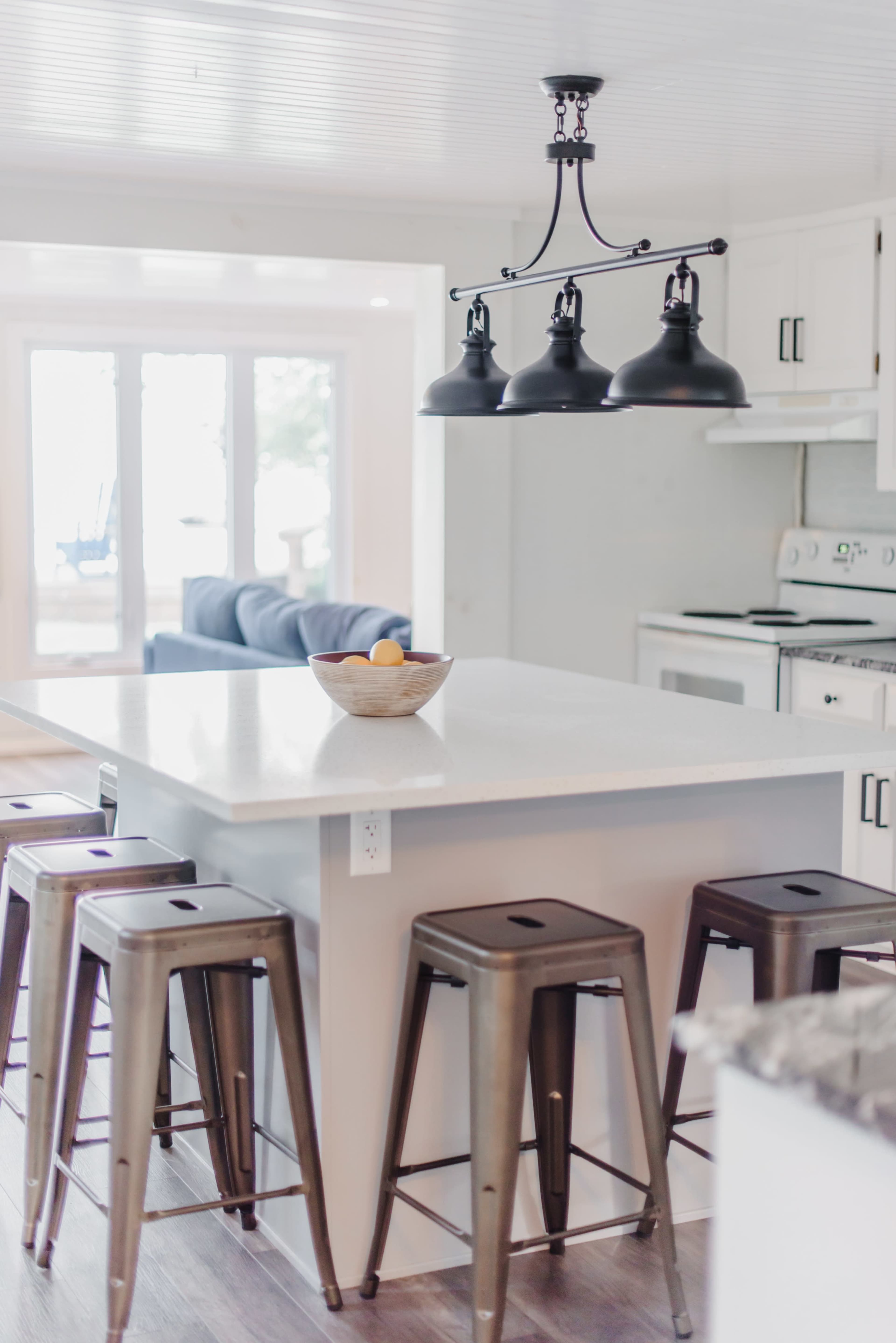 The image shows a modern kitchen with a white island featuring a bowl of fruit, surrounded by metal stools, and pendant lights overhead.