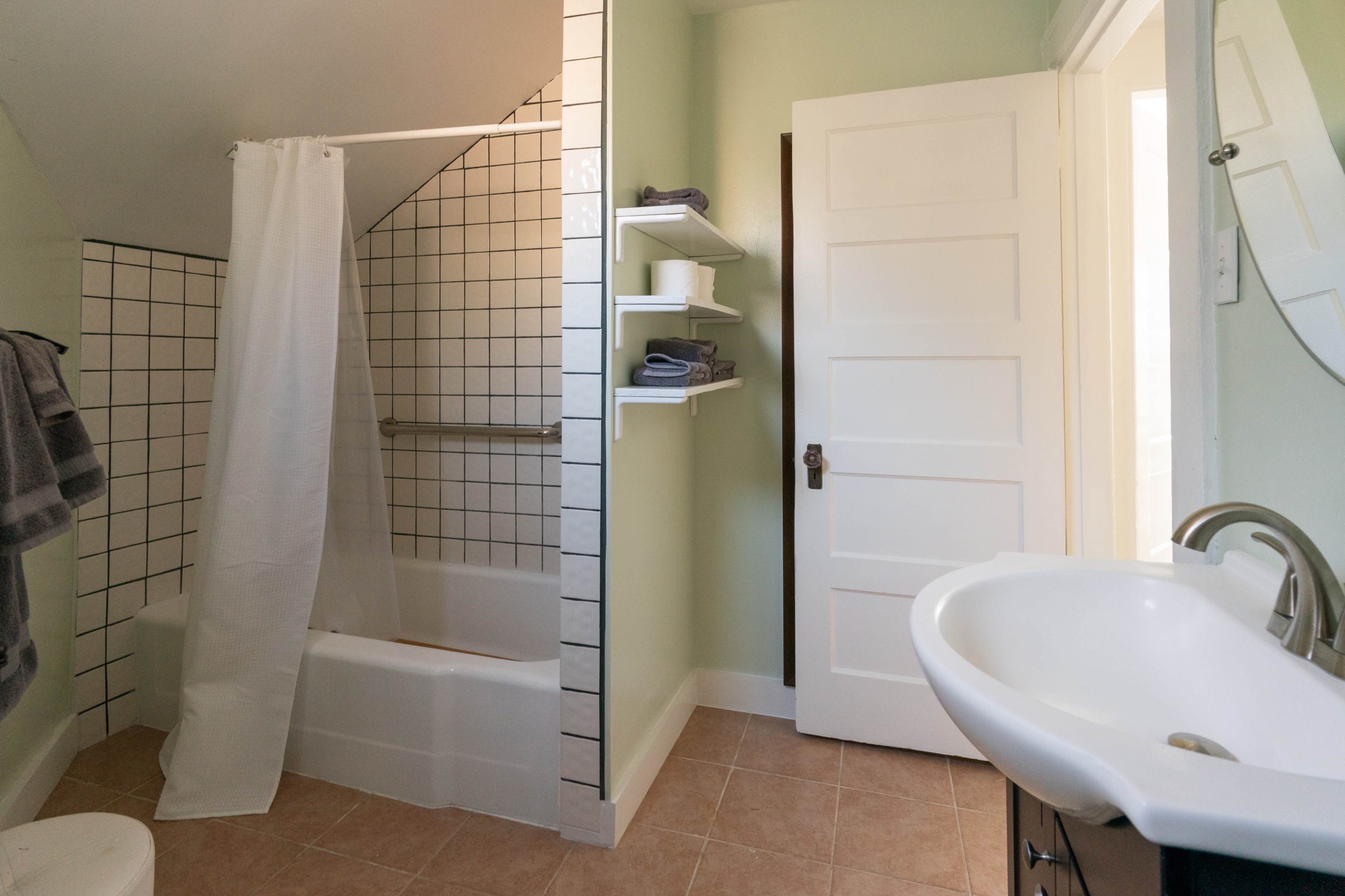 The image shows a bathroom featuring a shower/bath combination, shelves with towels, and a sink with a mirror against a light green wall.
