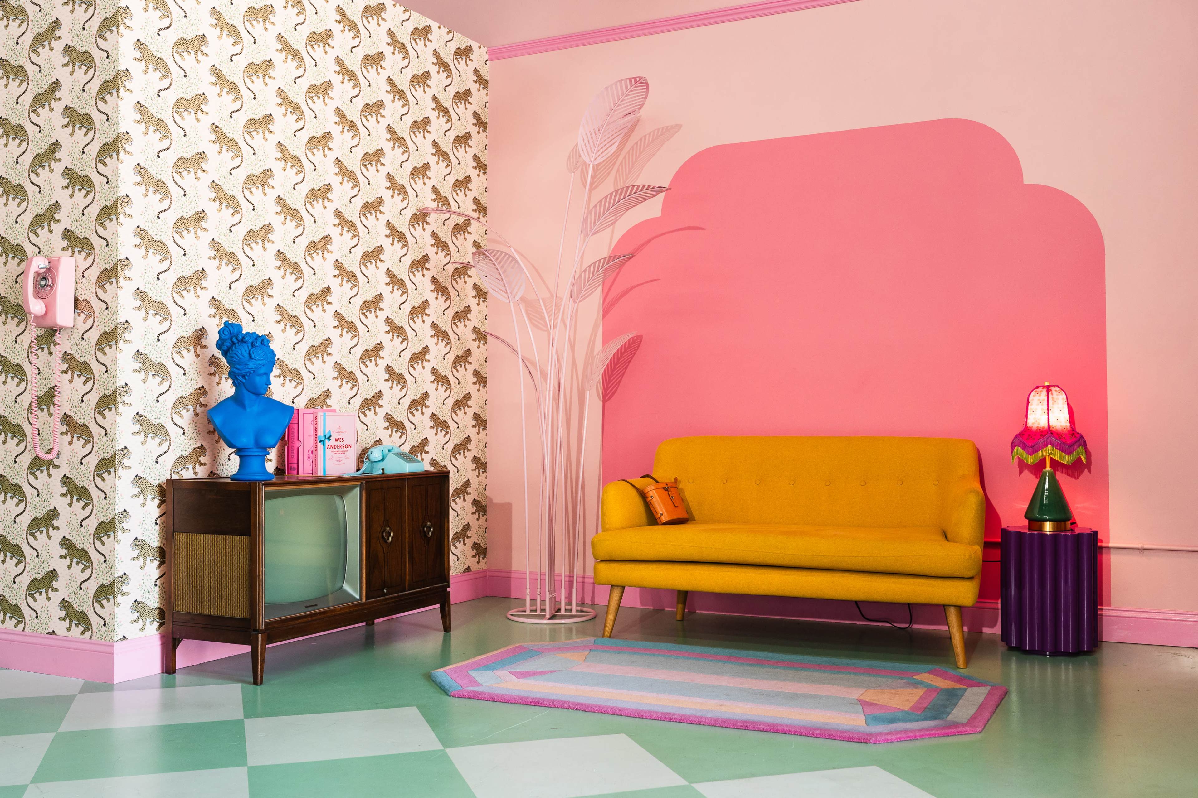 The scene features a brightly colored room with a yellow sofa, a retro television on a wooden cabinet, and decorative elements including a pink wall and patterned wallpaper.