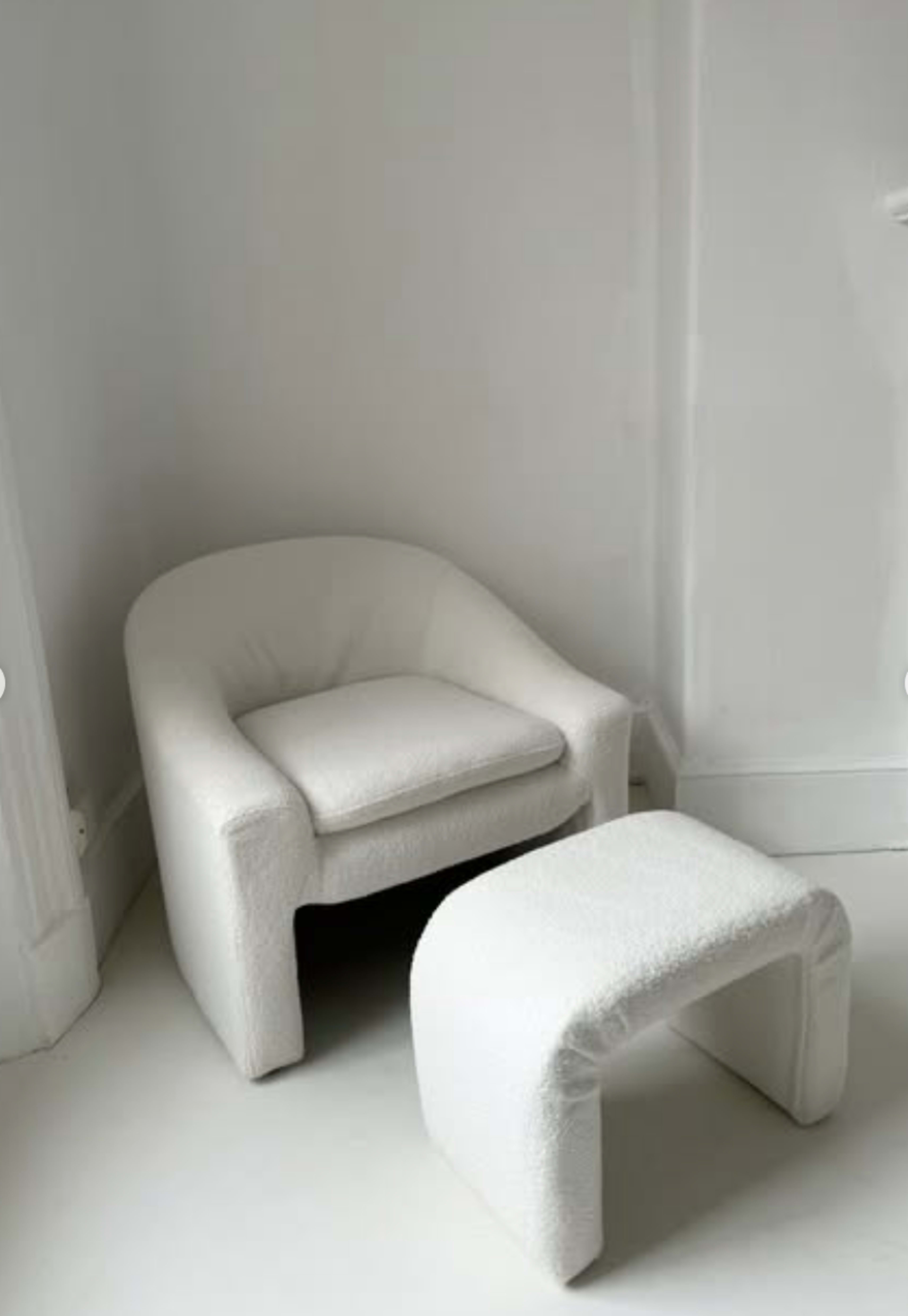 A white, textured armchair is positioned next to a matching small stool in a minimalist room with white walls.