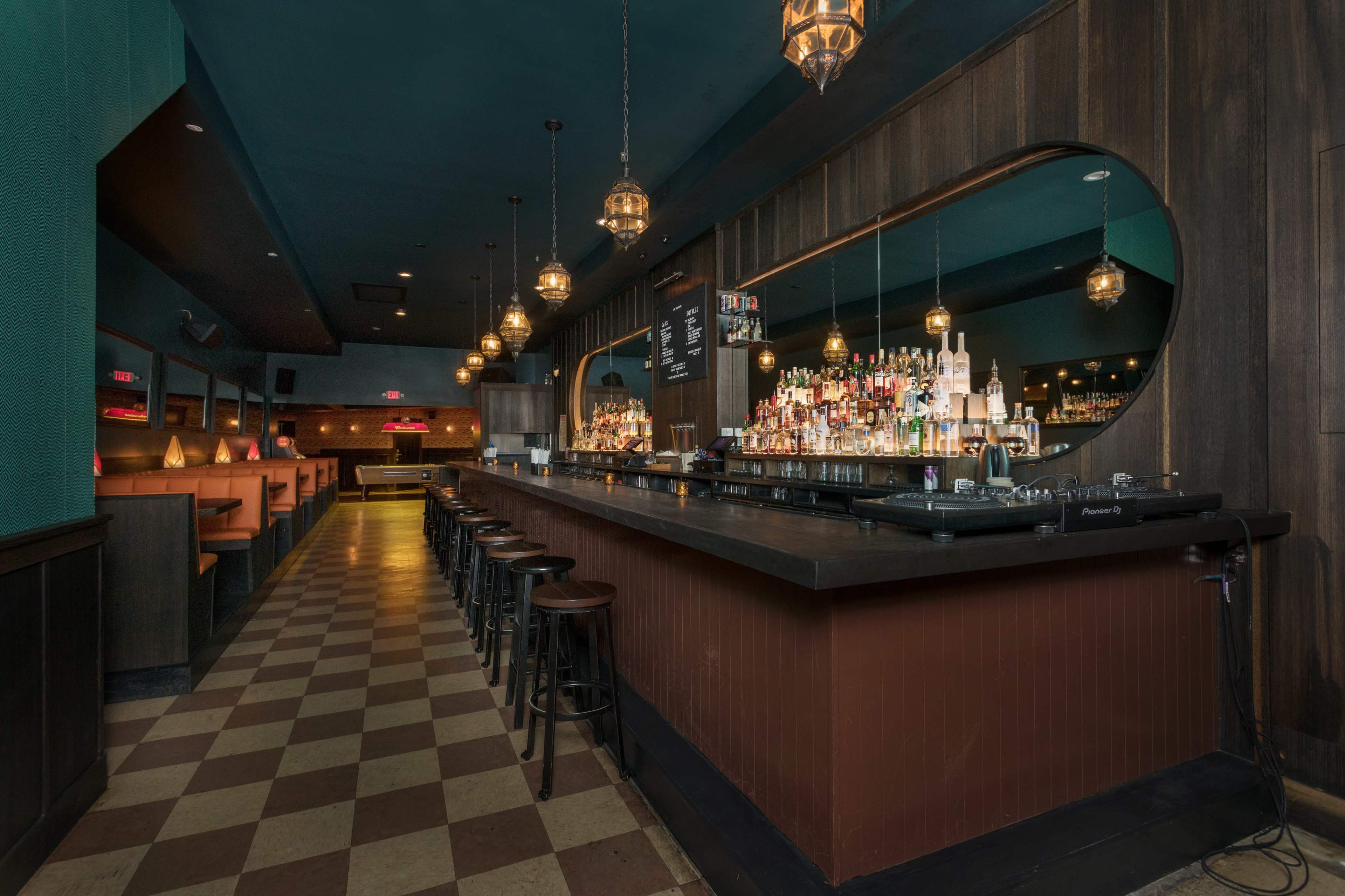 The image shows a dimly lit bar with a long countertop, lined with stools, and shelves filled with various bottles of liquor against a backdrop of dark wood paneling.