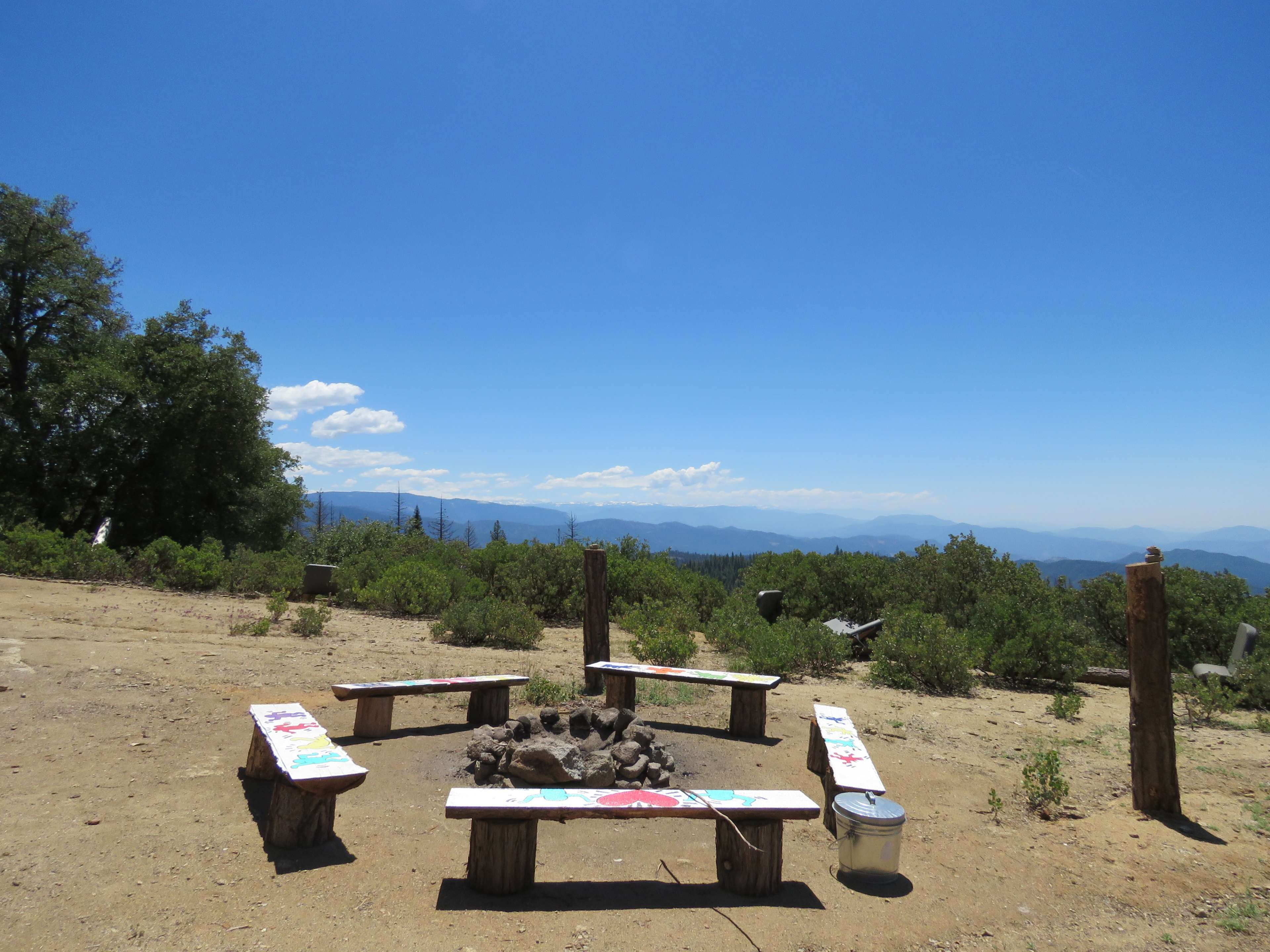 A campfire circle with benches around a fire pit, set against a clear blue sky and mountain landscape.