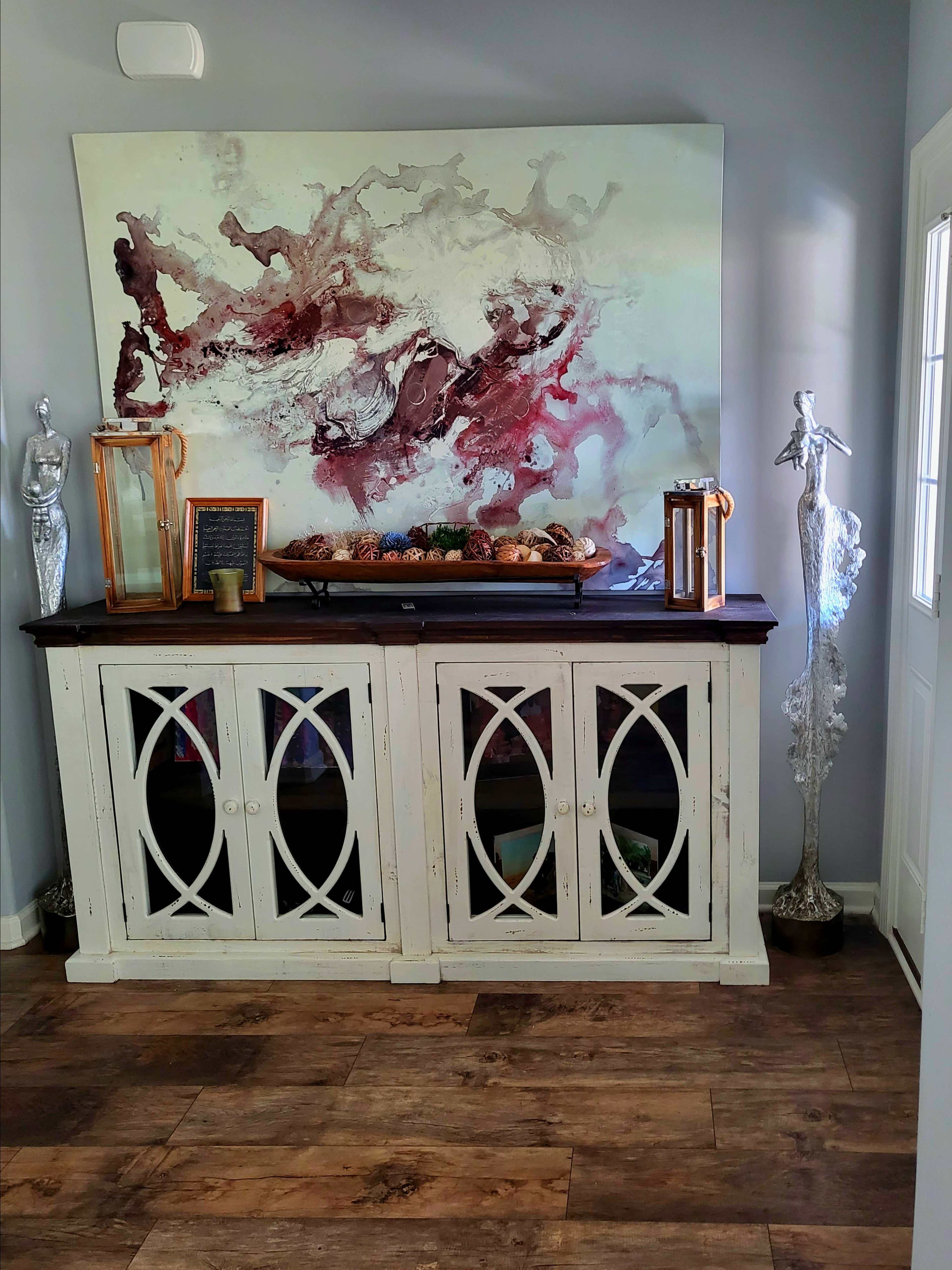 A white sideboard with glass doors displays decorative items, including candles and a large abstract painting, against a light gray wall.