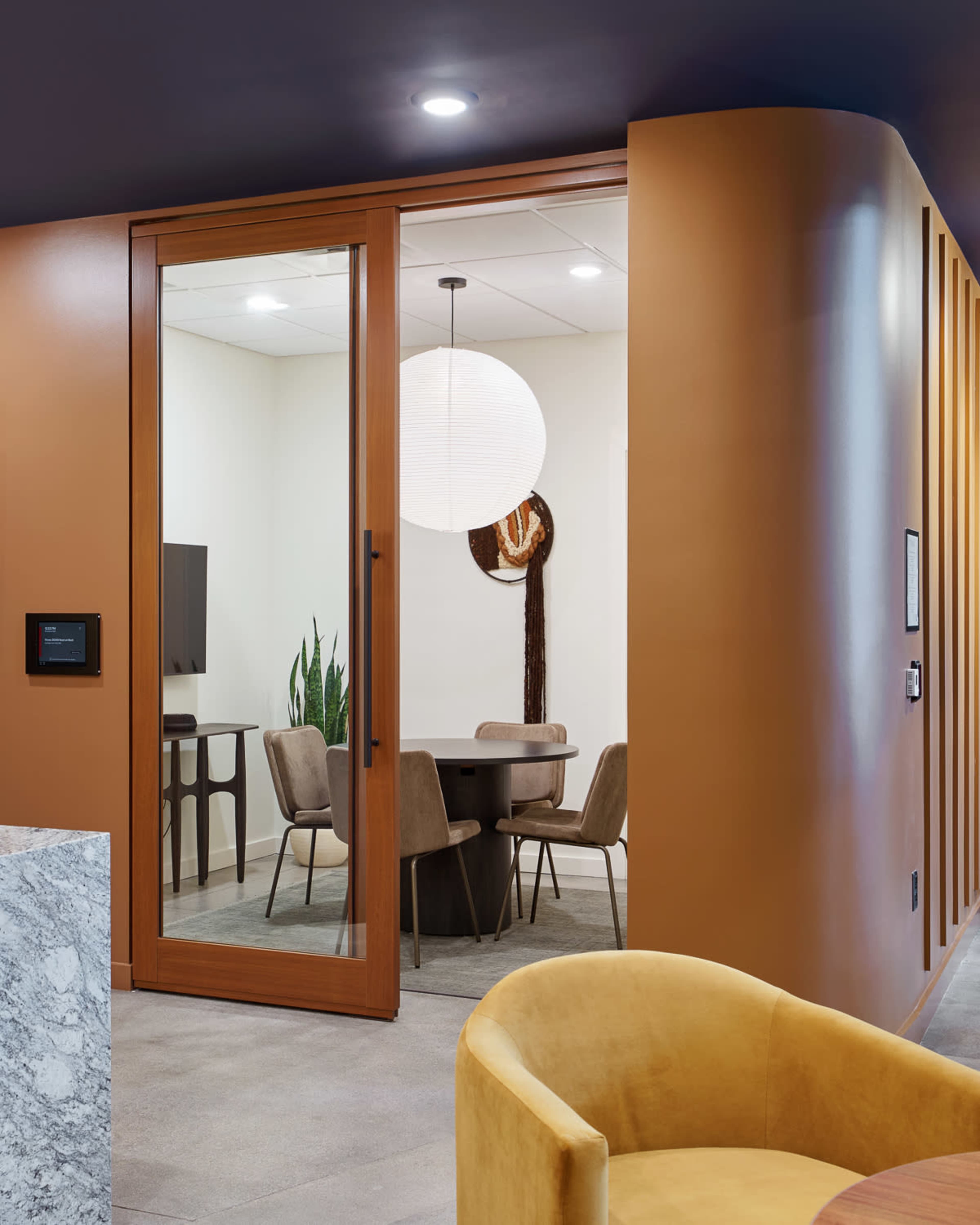 A modern conference room with a circular table and upholstered chairs is visible through a glass door, set against warm, orange-toned walls.