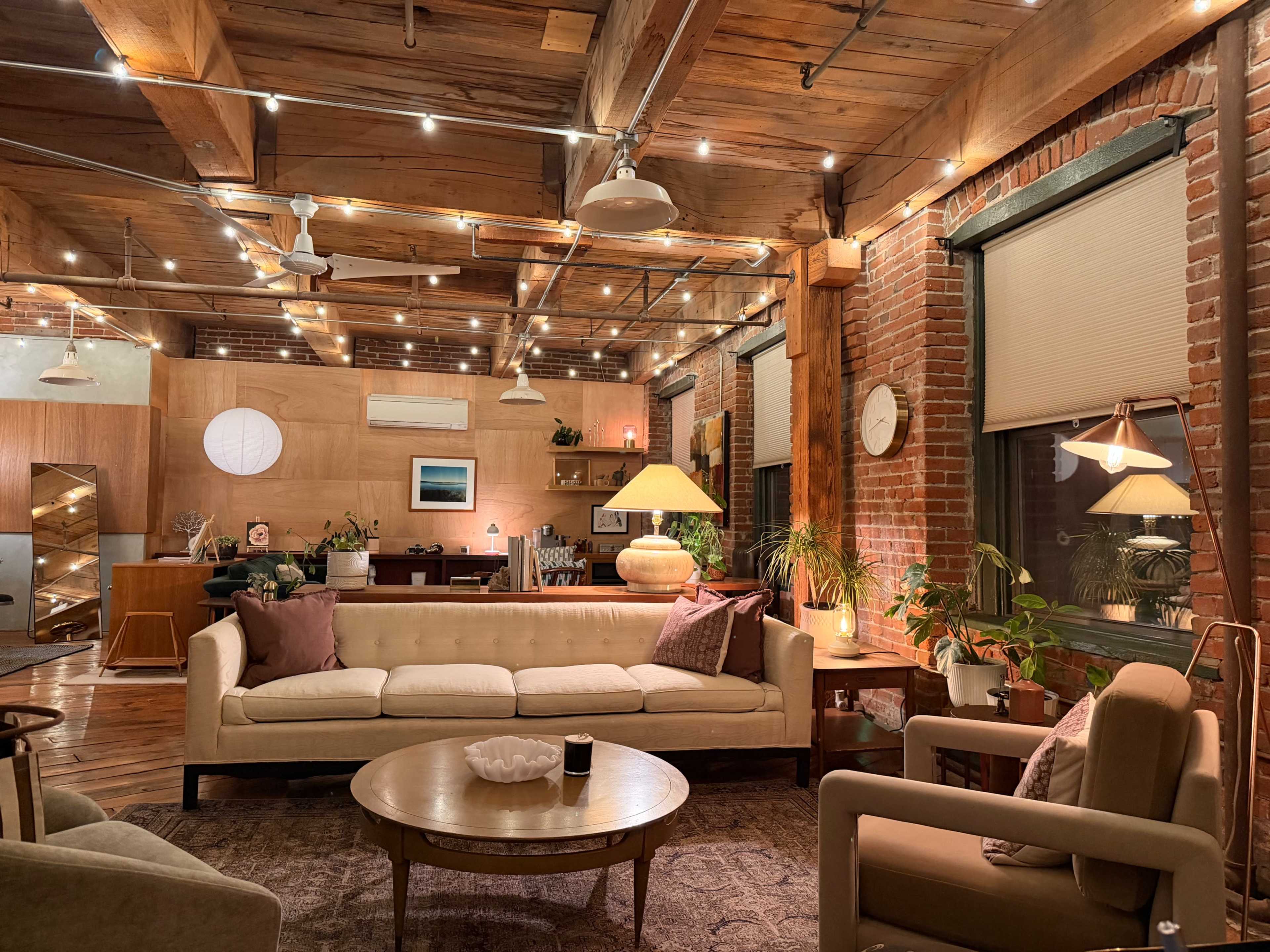 The image shows a cozy, well-lit living space with exposed wooden beams, brick walls, and comfortable furniture arranged around a central coffee table.