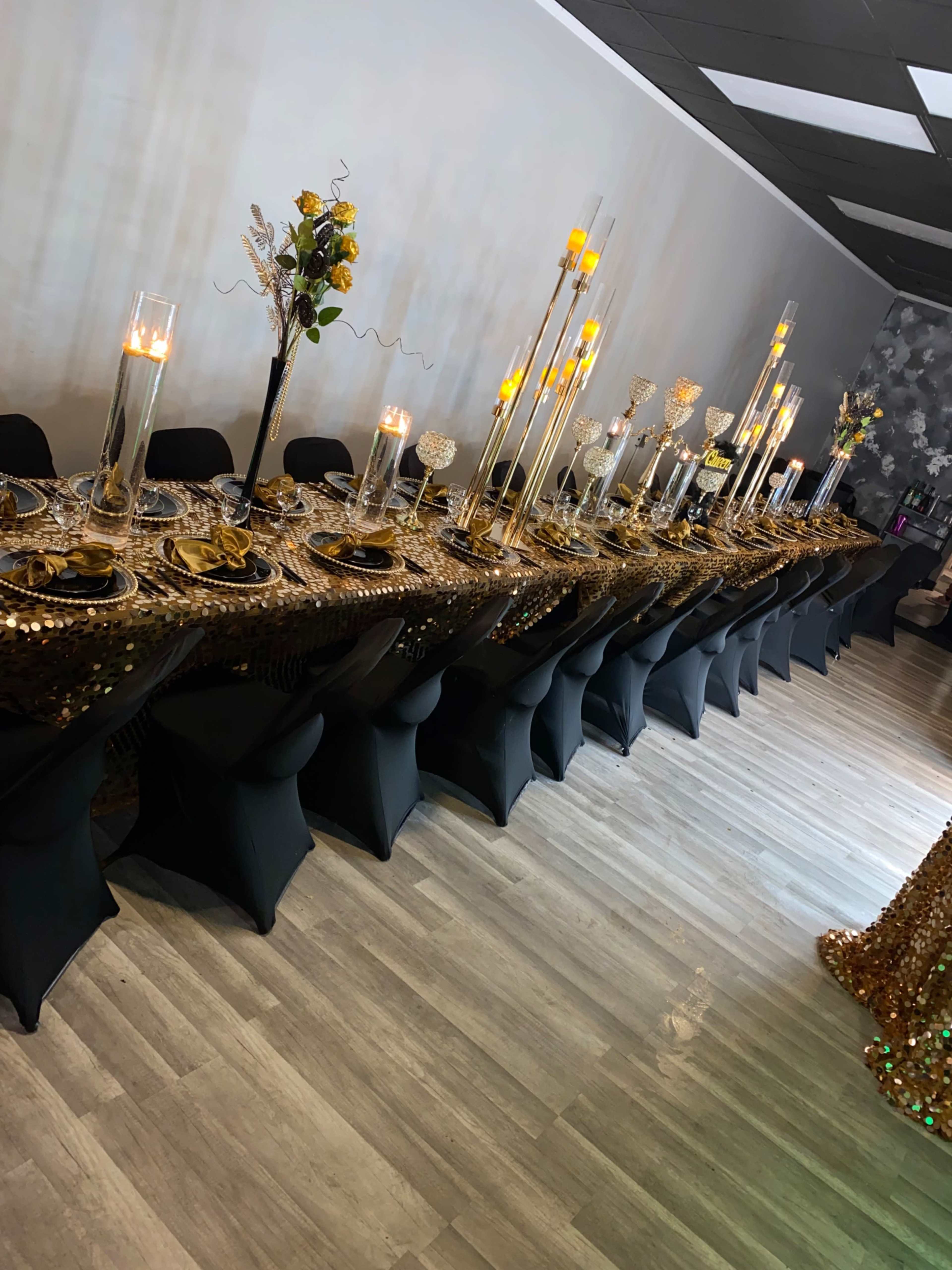 A long banquet table dressed in gold and black decor sits in a well-lit room, featuring elegant centerpieces and candle arrangements.