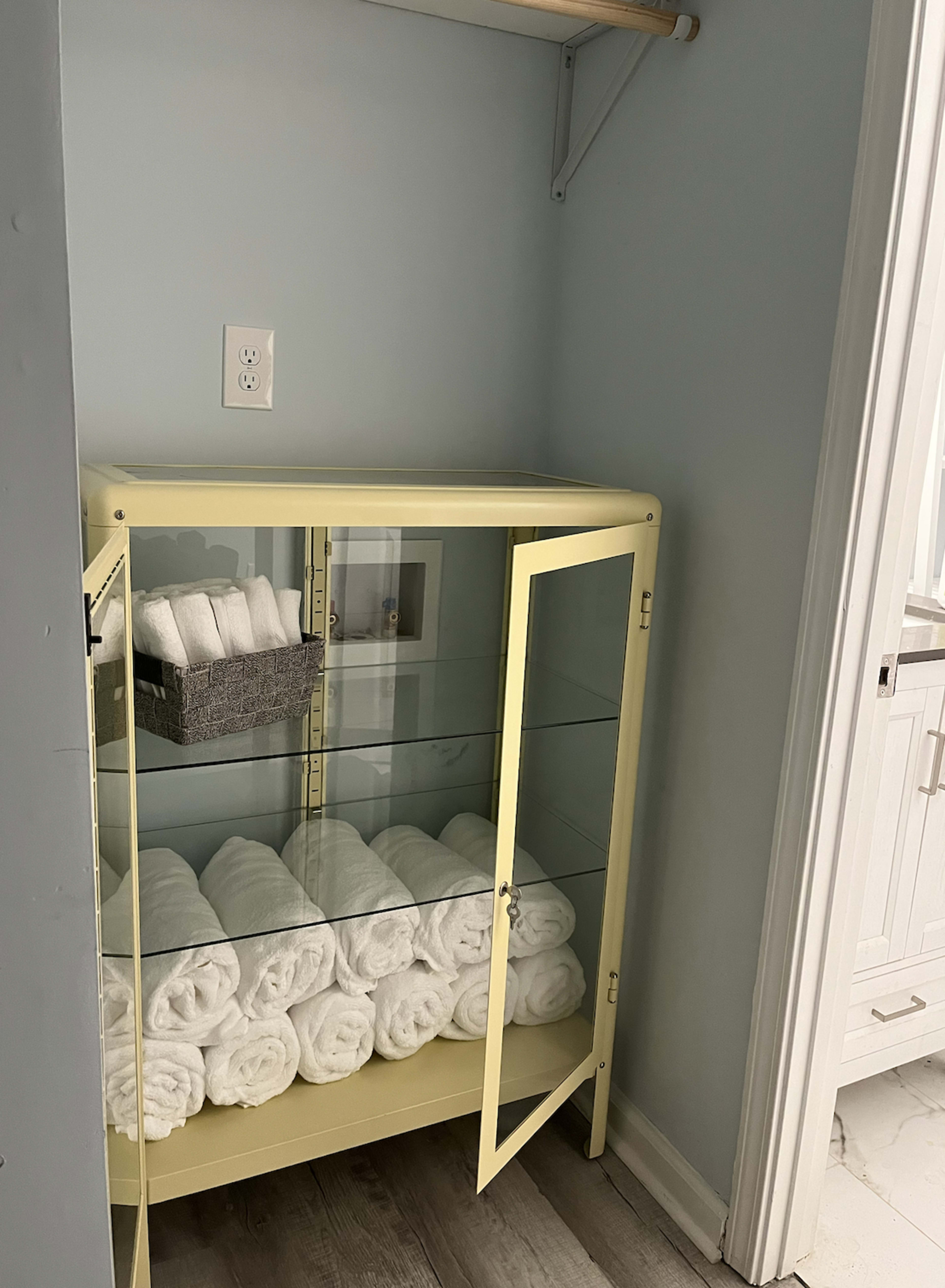 A yellow cabinet with glass doors displays neatly rolled white towels and a gray basket on a shelf.