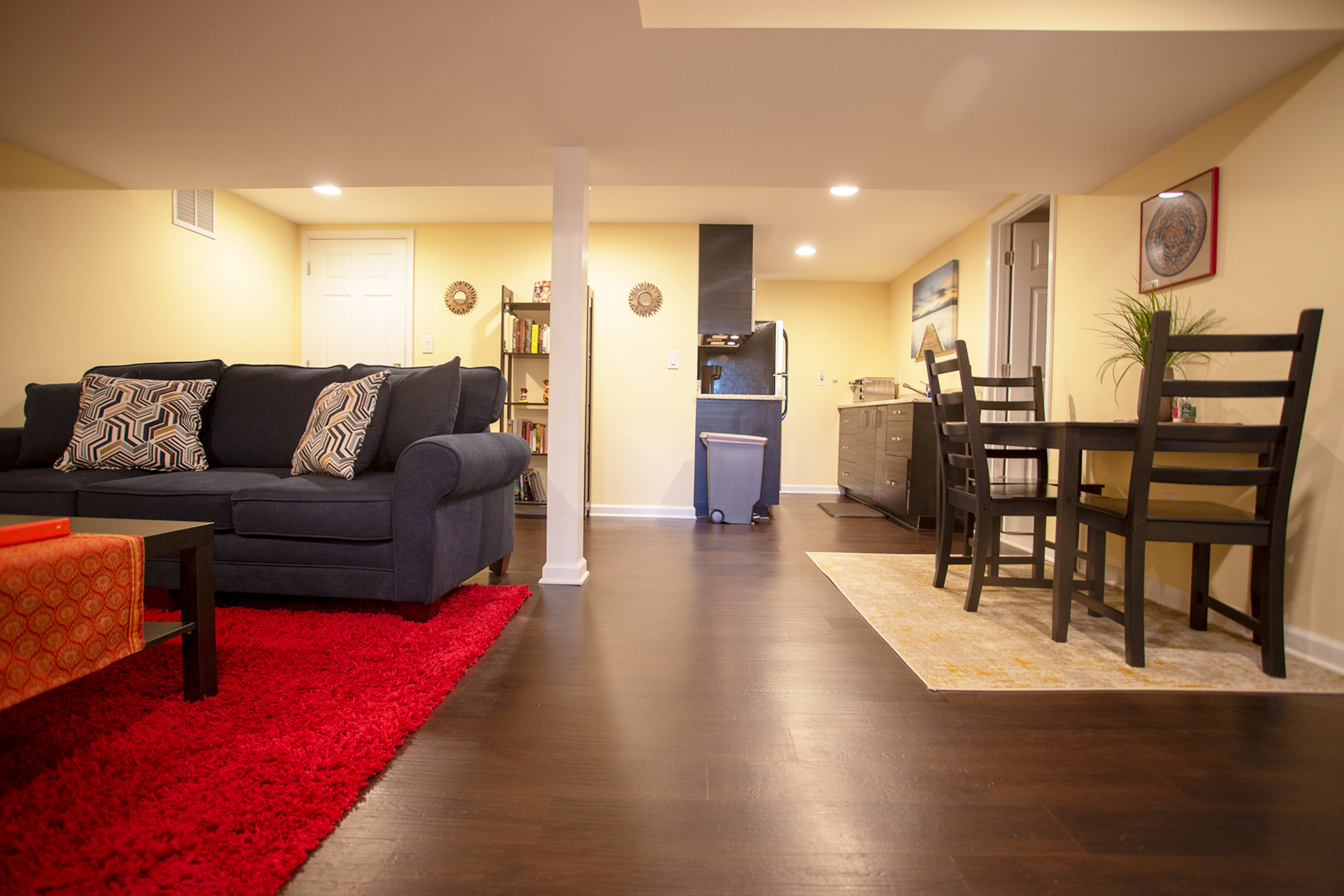 The image shows a well-lit basement living area featuring a sofa, dining set, and a kitchenette in a cozy layout.