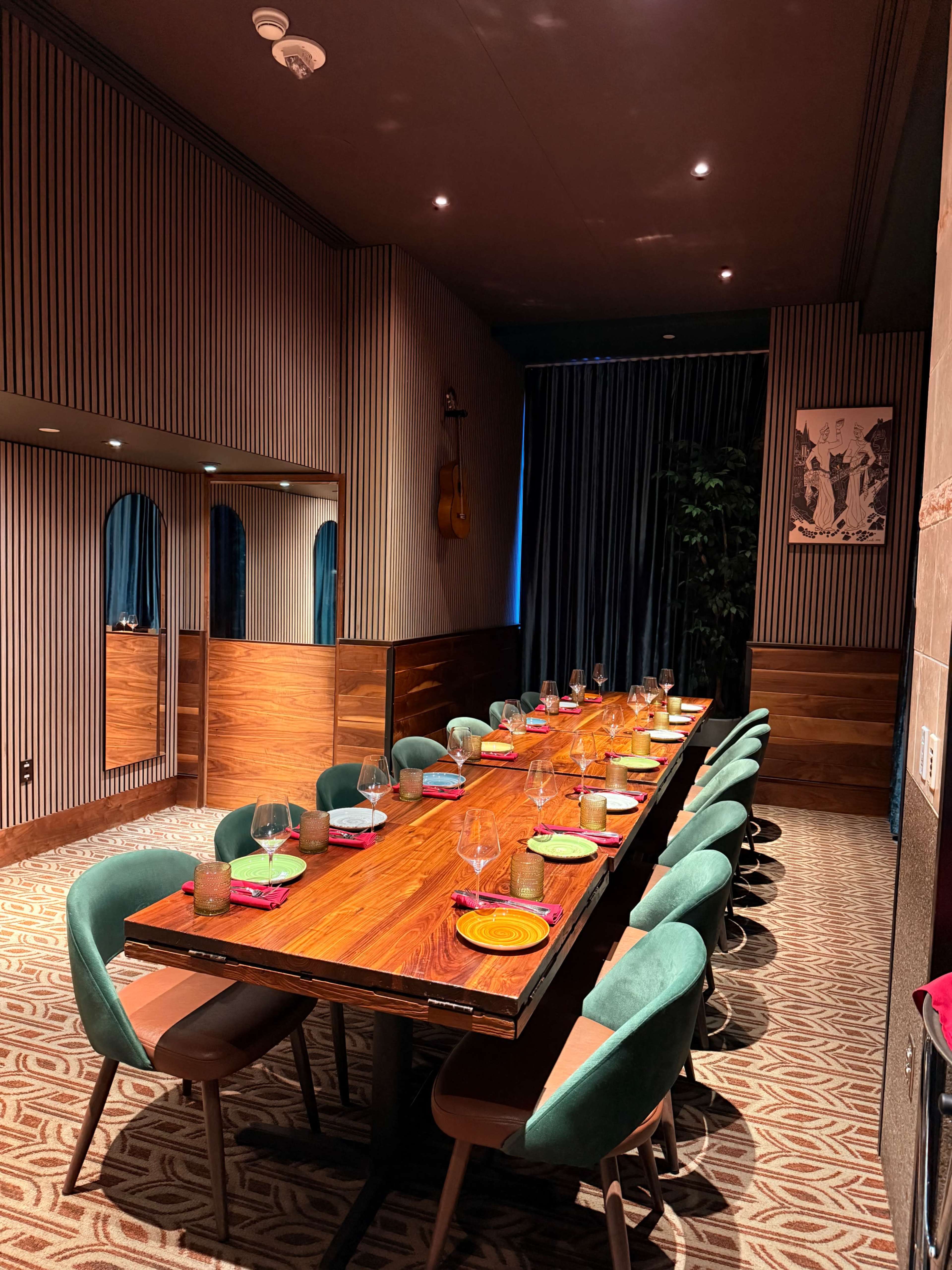 A long wooden dining table is set with colorful plates and glasses in a modern, stylish restaurant room featuring a striped wall and blue curtains.