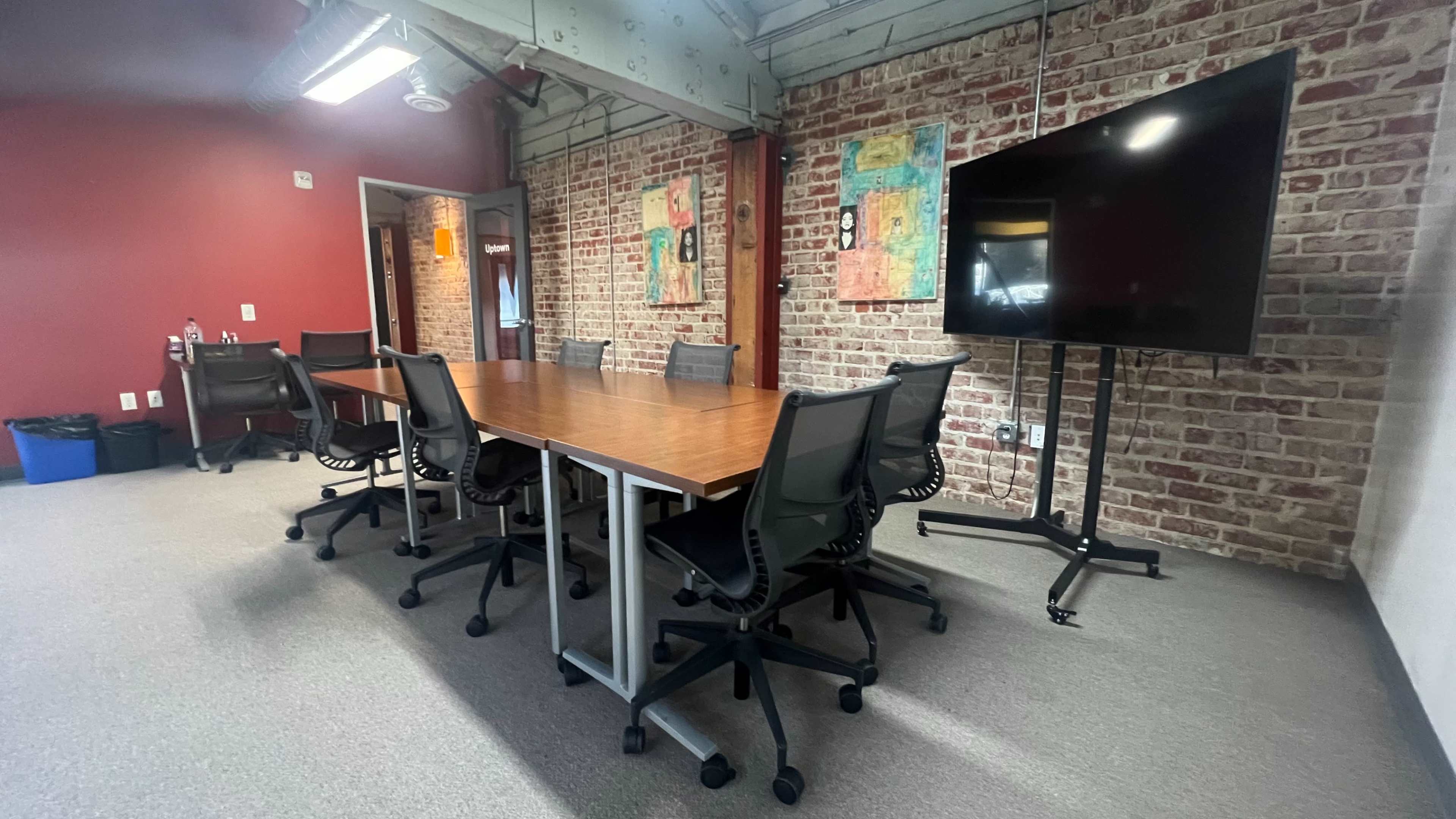 A conference room features a large wooden table surrounded by black office chairs, with exposed brick walls and a large screen on a stand.