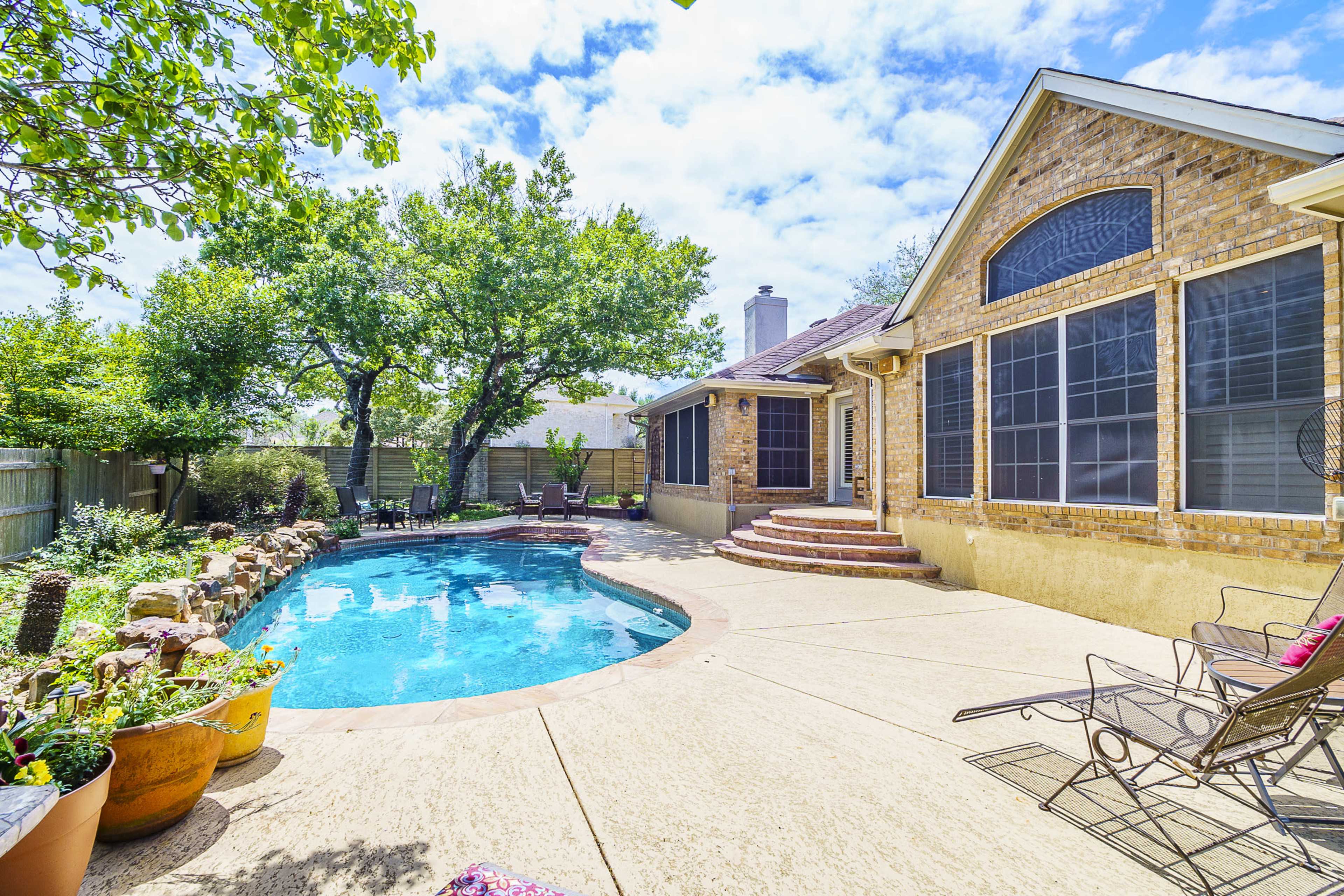A backyard features a swimming pool surrounded by concrete, with a house partially visible on one side and lush greenery nearby.