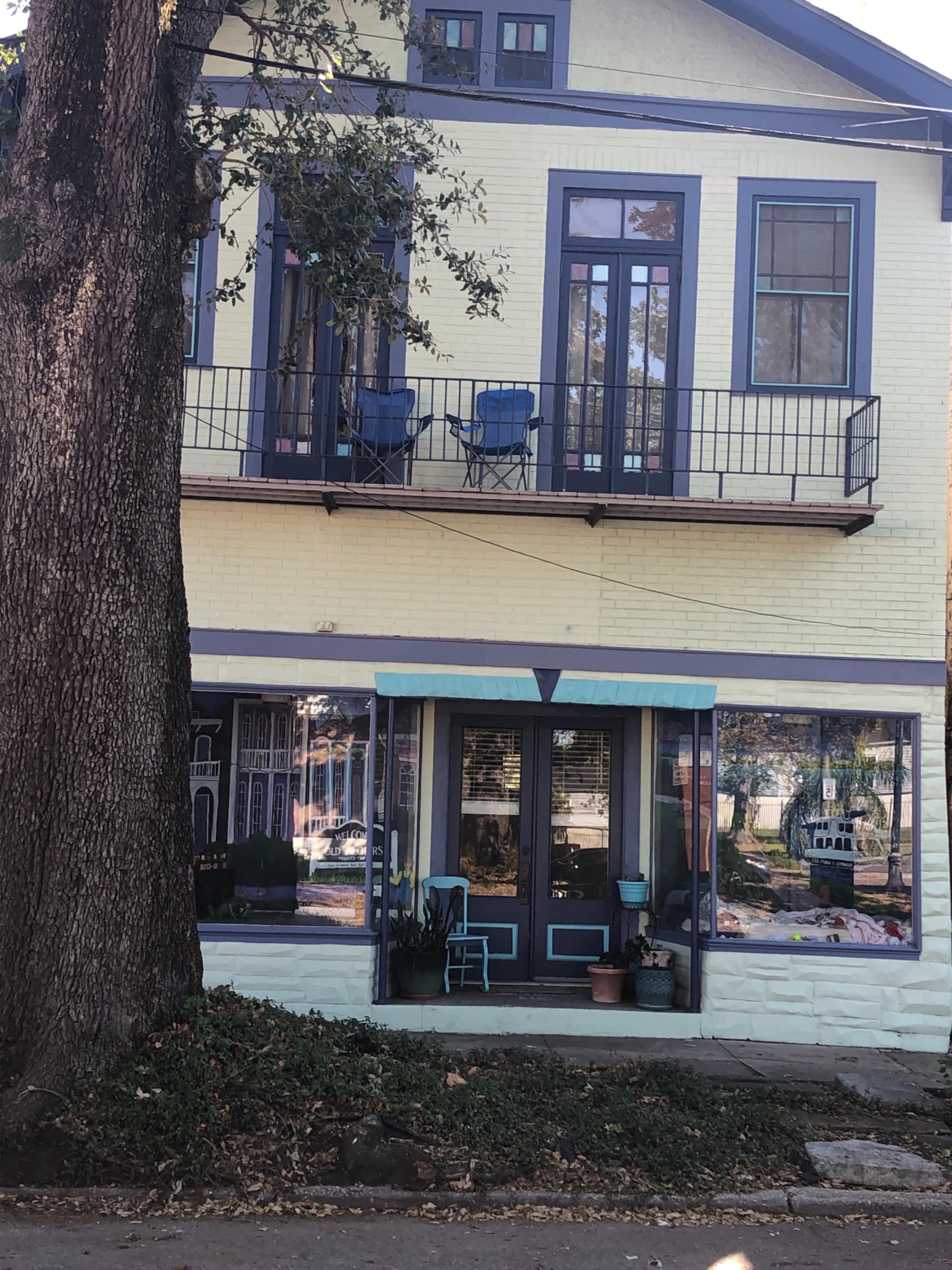 A yellow two-story building with a balcony, blue chairs, and storefront windows displaying various items on the ground floor.