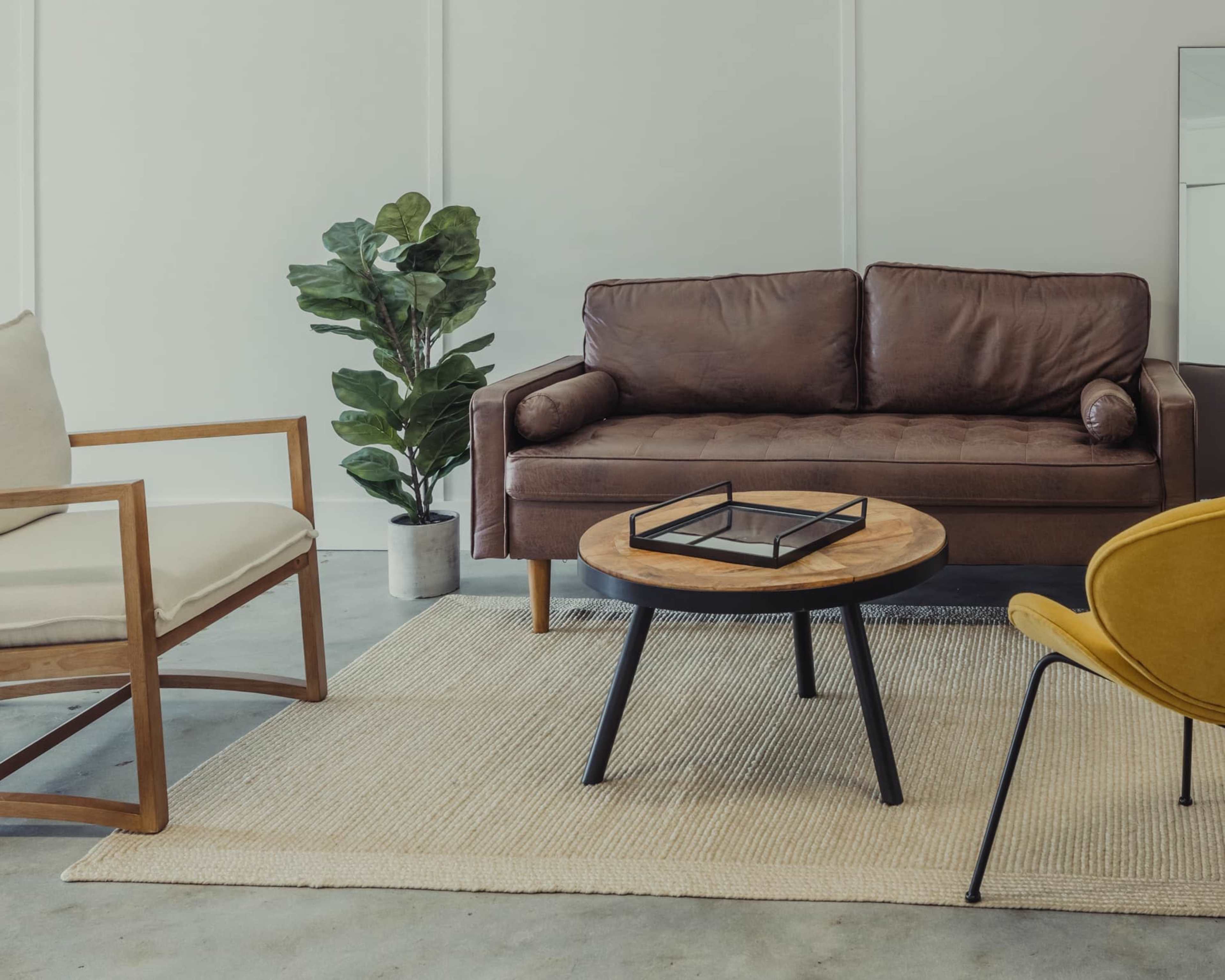 A cozy living room setting with a brown leather sofa, a round wooden coffee table, a light-colored area rug, and a potted plant.