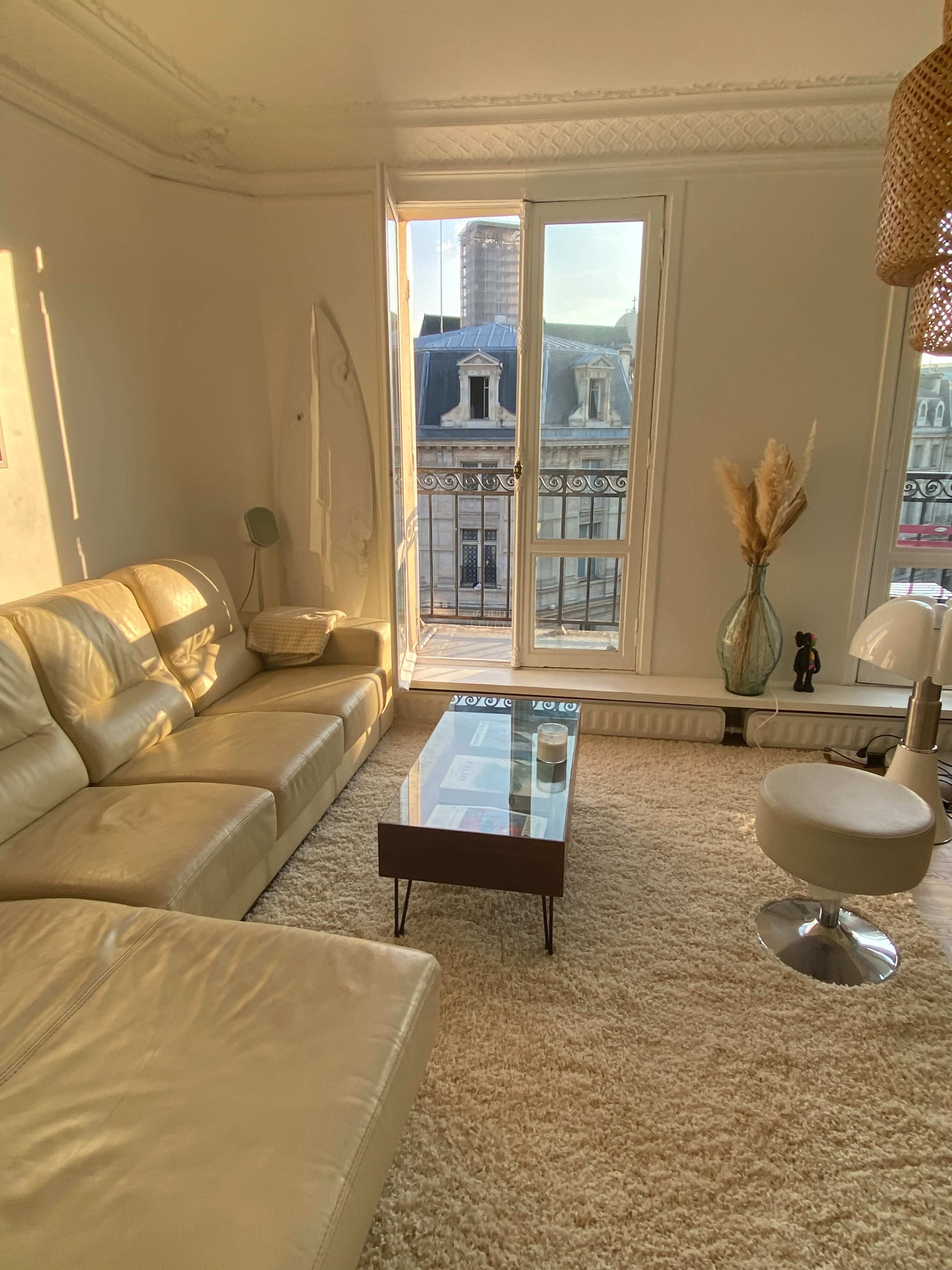 A cozy living room features a beige leather sectional sofa, a glass coffee table, and large windows overlooking a cityscape.