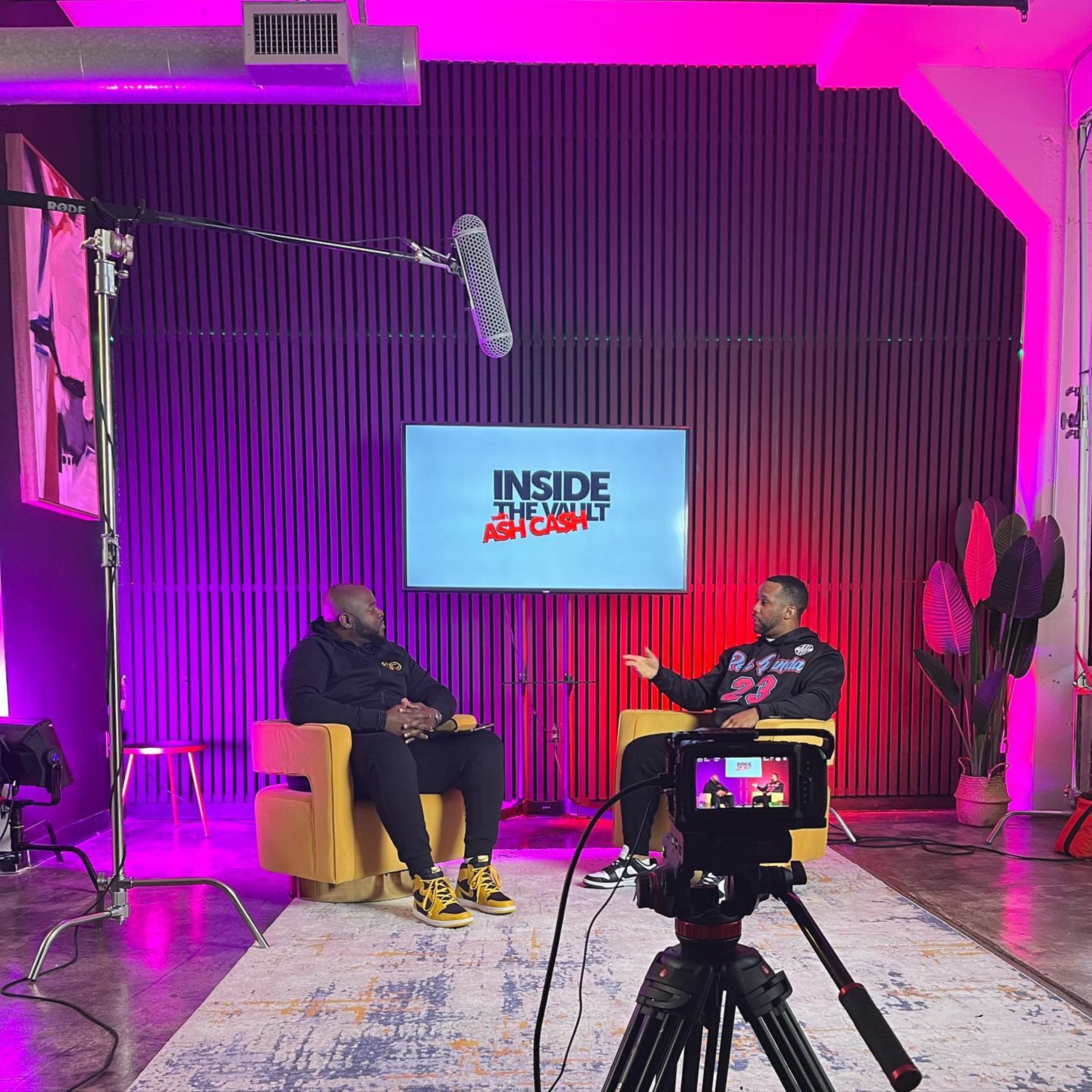 The image shows two men sitting in yellow chairs in a studio setting, with a large screen in the background displaying the text "INSIDE THE VAULT" and colorful lighting.