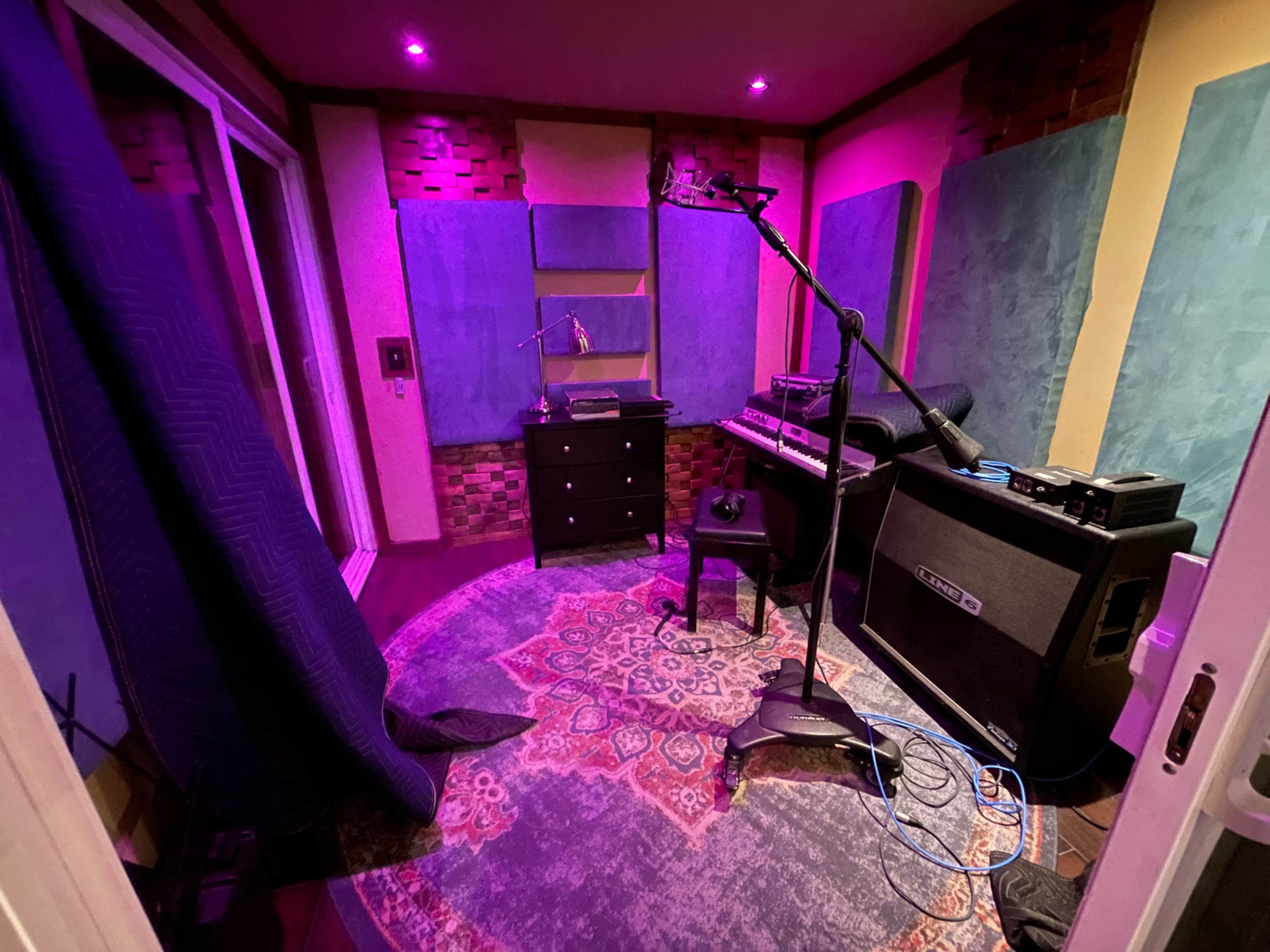 The image shows a sound recording studio with acoustic panels on the walls, a microphone on a stand, and various musical equipment.
