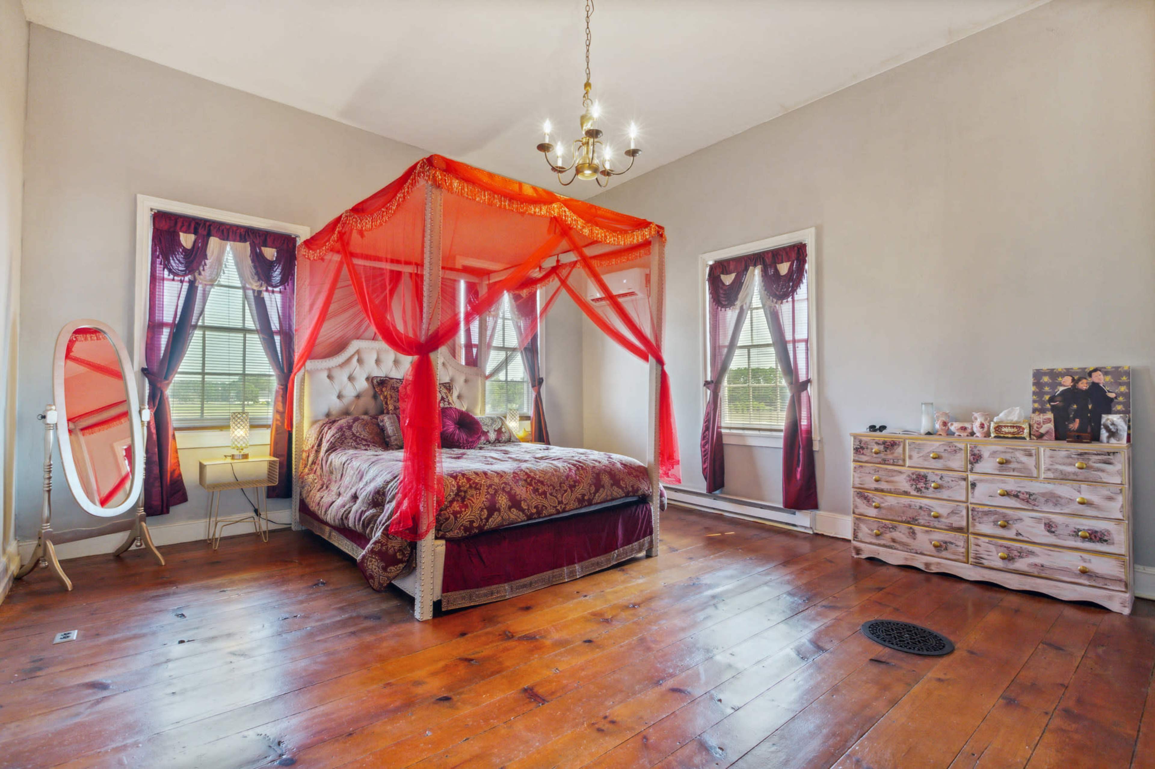 The image shows a bedroom featuring a four-poster bed draped with red fabric, two large windows with curtains, and a vintage dresser.