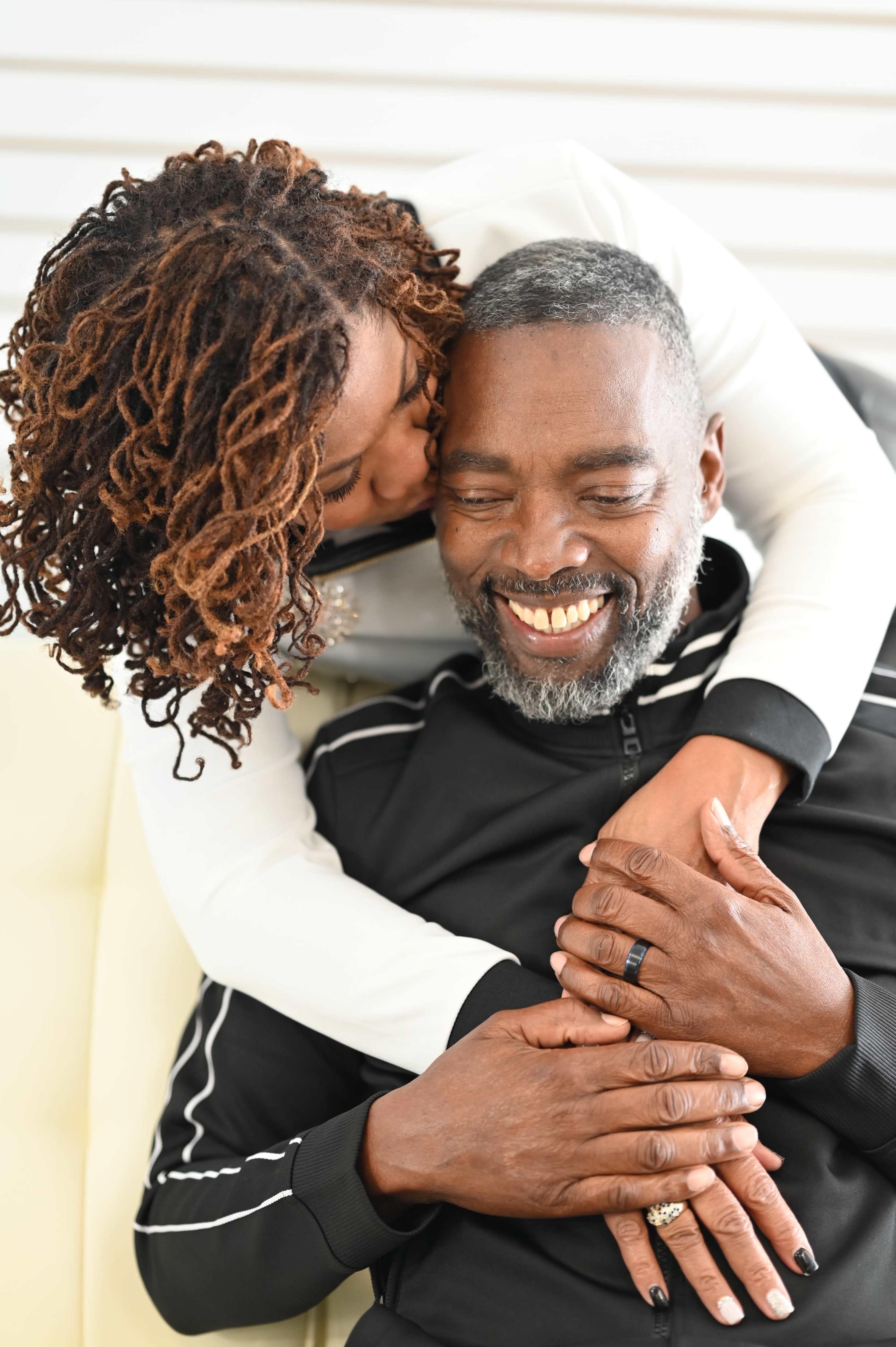 A man and a woman embrace on a couch, with the woman leaning in and kissing the man on the cheek while he smiles.