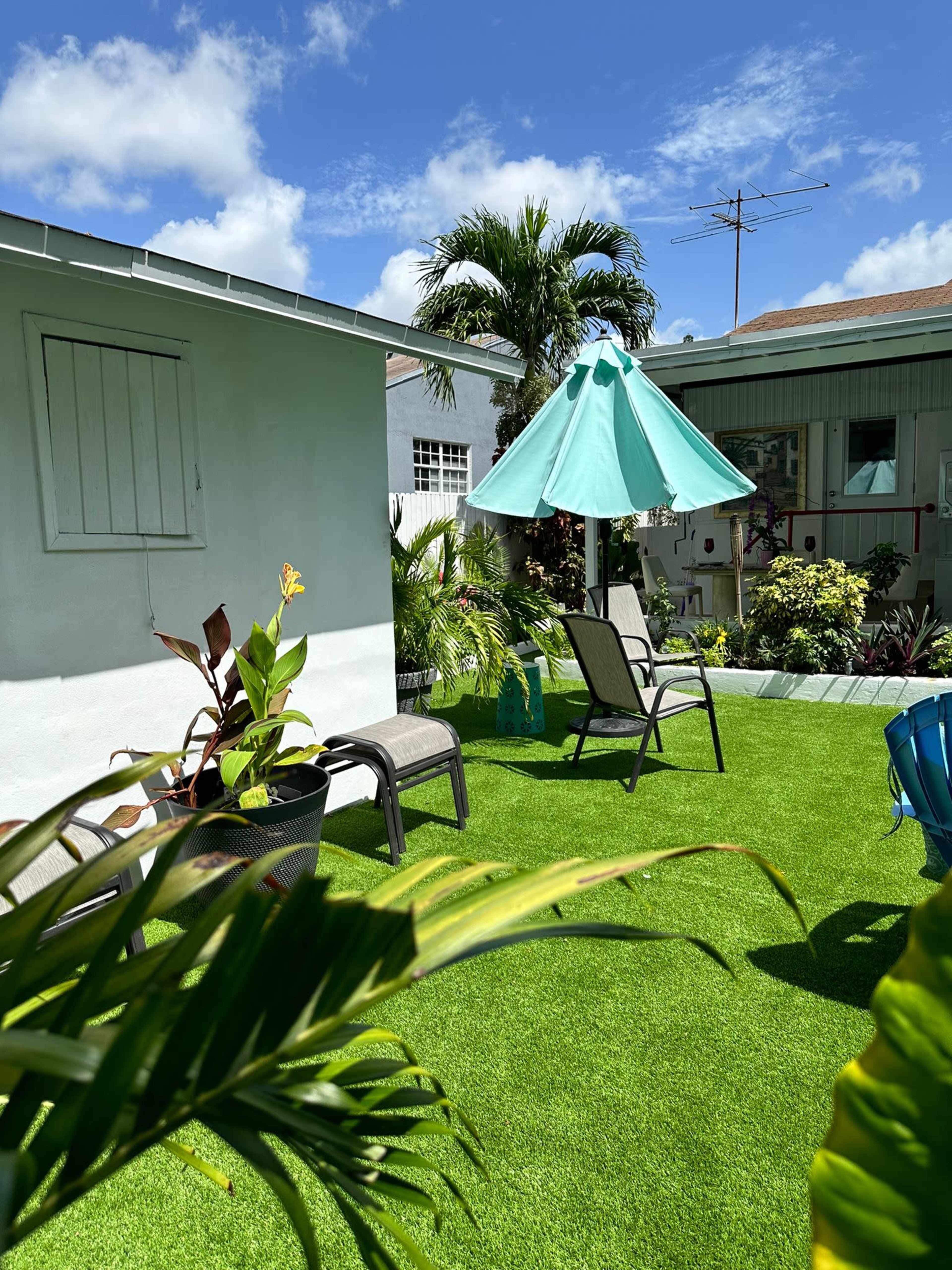 The image shows a landscaped backyard with artificial grass, a blue umbrella over a seating area, and tropical plants surrounding the space.
