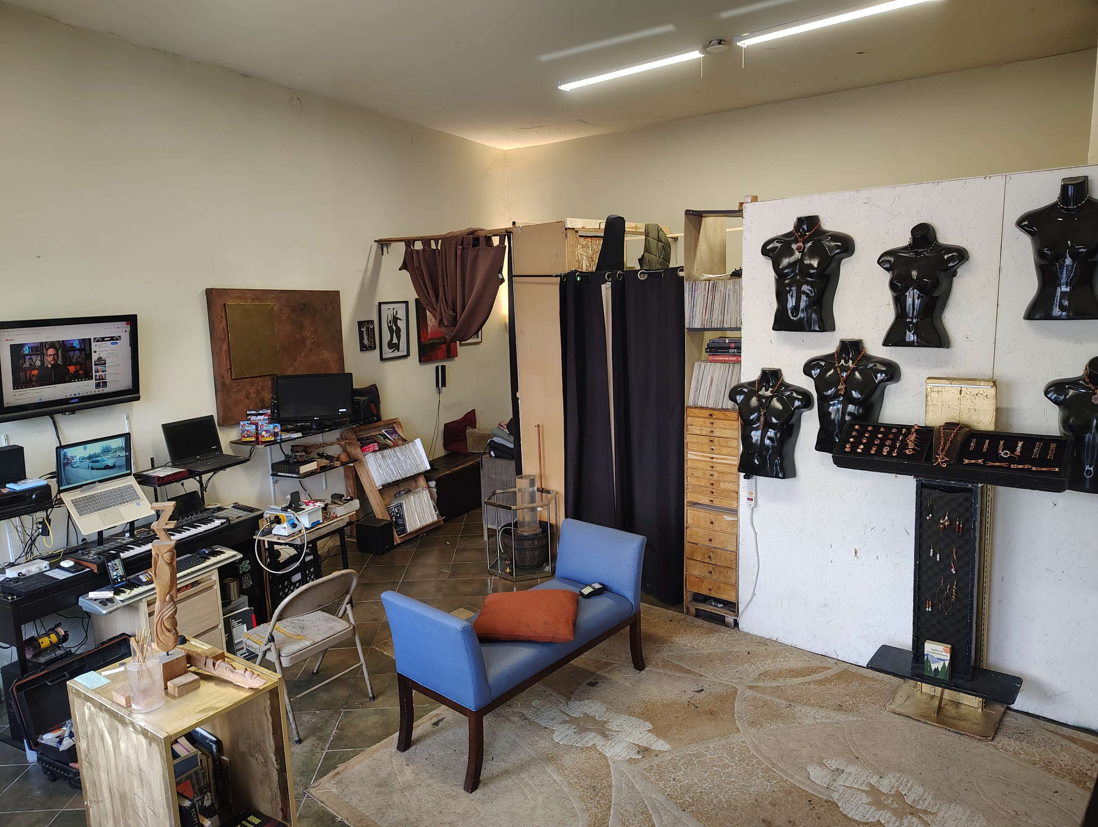 The image shows an artist's studio featuring a blue chair, a computer setup, various tools and materials, and display mannequins on the walls.